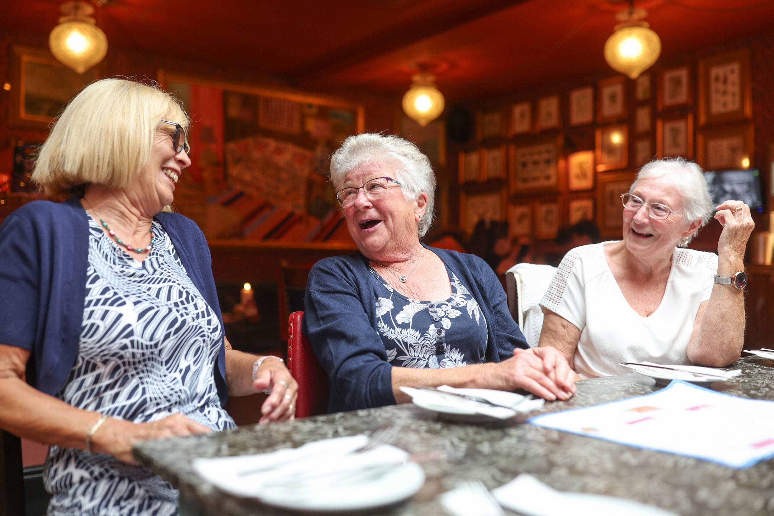 Three older women sit together at an Antico restaurant table, laughing and enjoying each other's company. Plates and menus are on the table, and the background is warmly lit with framed pictures on the walls.