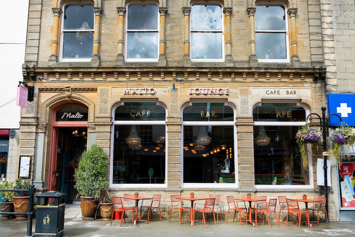 A stone-fronted café and bar named "Matto Lounge" with large windows, outdoor seating with orange chairs and tables, potted plants, and hanging flower baskets on a city street offers Malto-inspired drinks in a vibrant setting.