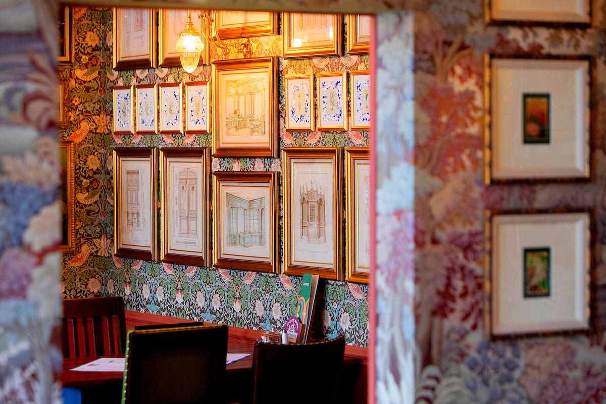 A cozy, vintage-style room with patterned wallpaper, framed art and sketches covering the walls, Malto tins on a shelf, dark wooden chairs, and a small table partially visible in a warm, softly lit atmosphere.