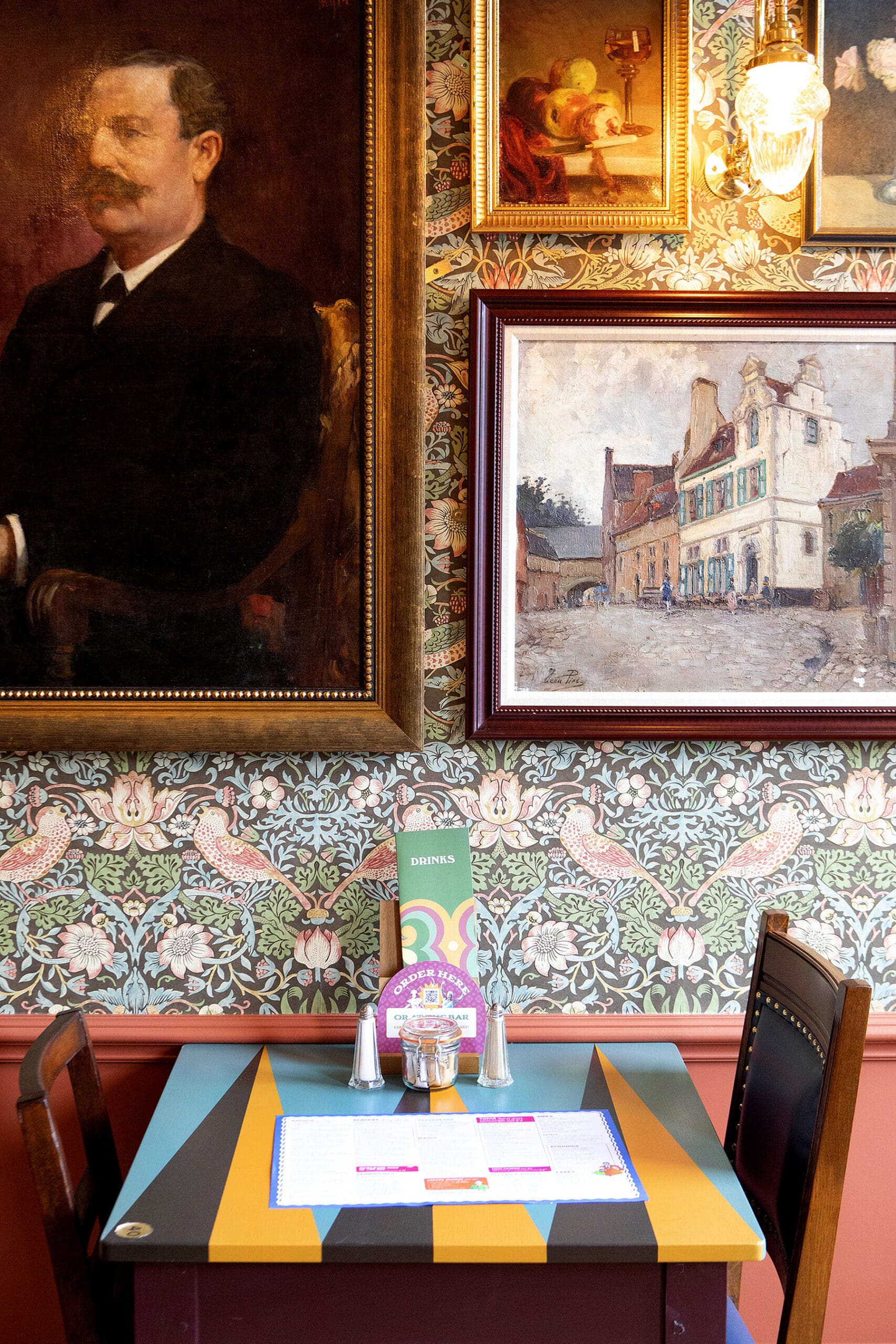 A small, colorful Malto café table for two is set with menus and condiments, against a wallpapered wall decorated with framed paintings, including a man’s portrait and a European street scene.