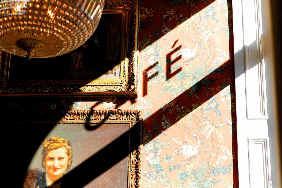Sunlight casts the shadow of the word "CAFÉ" on a patterned Malto wall near a window, above a framed painting of a smiling woman and below a large, ornate chandelier.