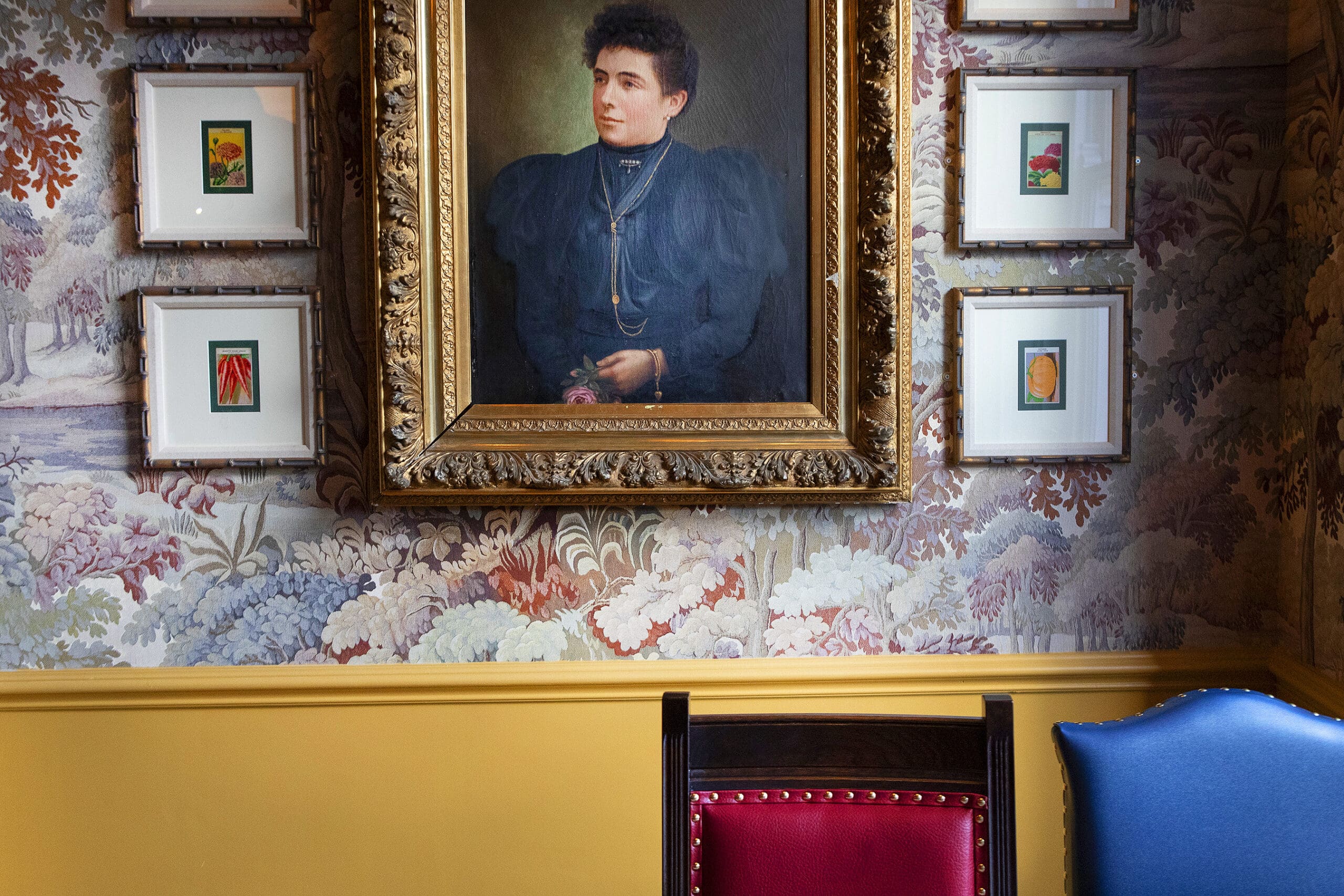A framed portrait of a woman in Victorian-era clothing hangs on a decorative floral Malto wallpapered wall, surrounded by small framed artworks. Below, a red and a blue chair sit against a mustard yellow wainscoting.