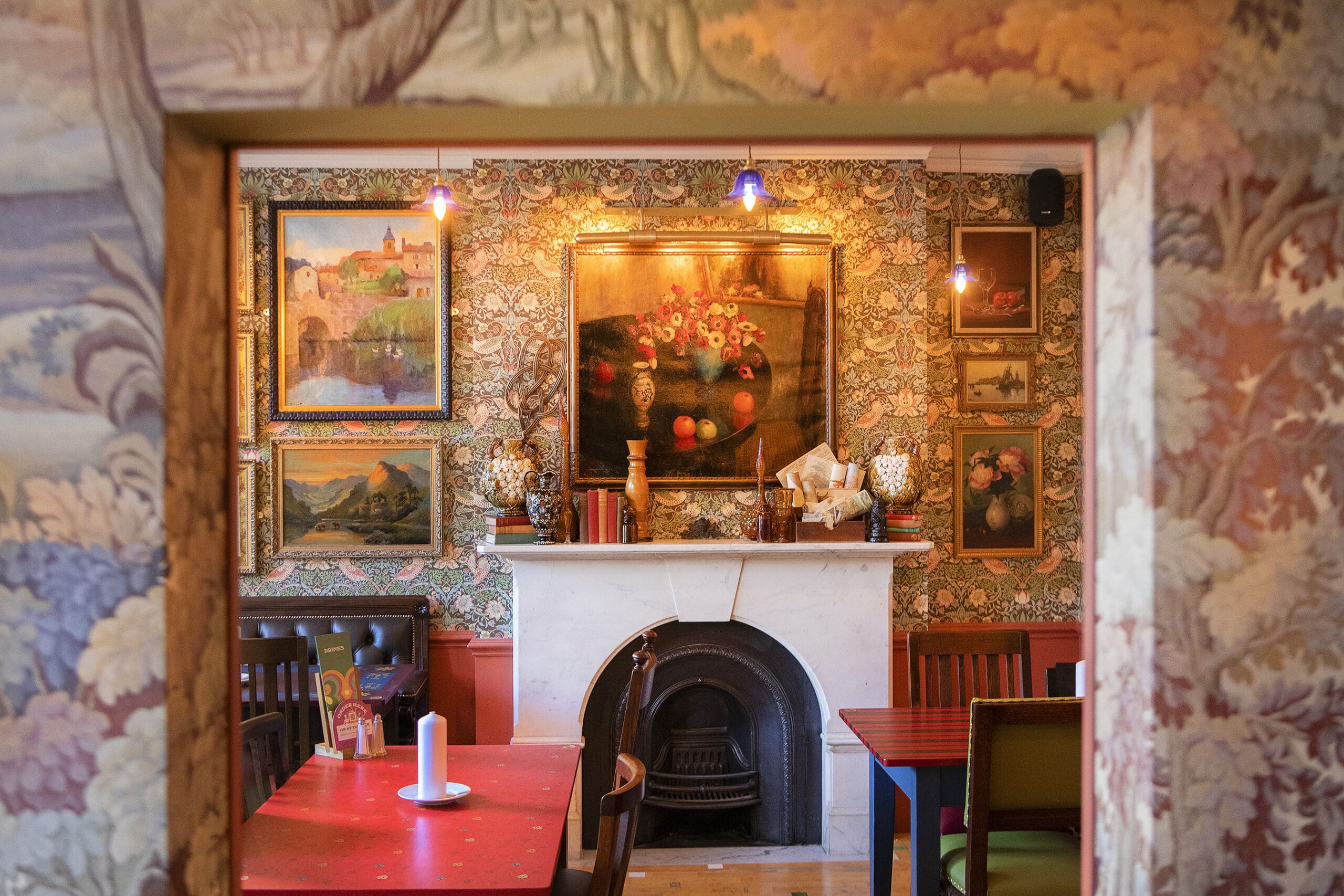 A cozy, vintage-style room with patterned wallpaper, a white marble fireplace, framed paintings, and a red table with chairs. Warm lighting and ornate decor enhance the inviting atmosphere—perfect for enjoying a Malto in comfort.