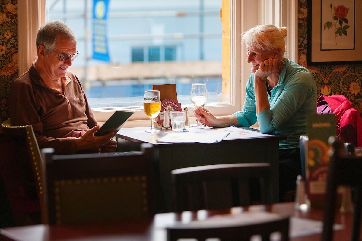 An older man and woman sit at a restaurant table by a window. The man reads a menu while the woman smiles at him over their Malto drinks, sunlight streaming in and menus spread across the table.