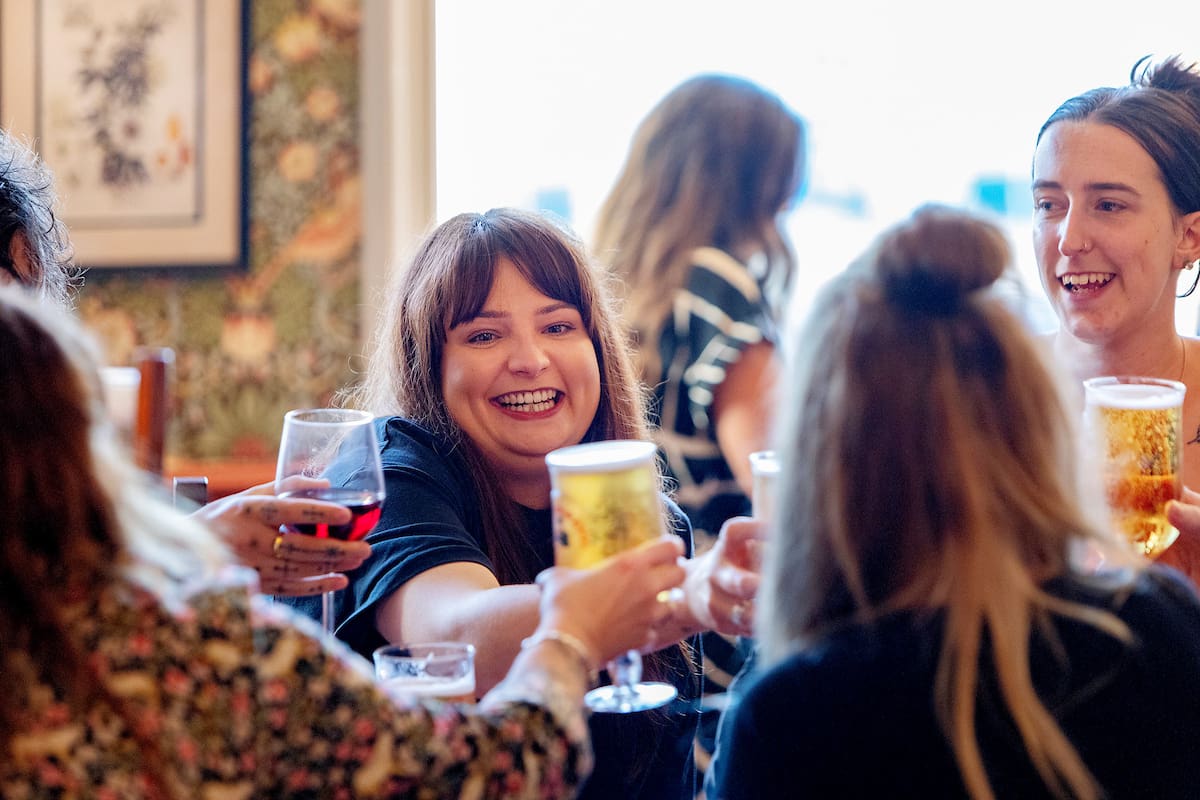 A group of women smiling and raising their glasses of Malto in a toast at a social gathering. One woman in the center, with brown hair and bangs, is laughing and reaching out with her drink. The atmosphere appears warm and joyful.