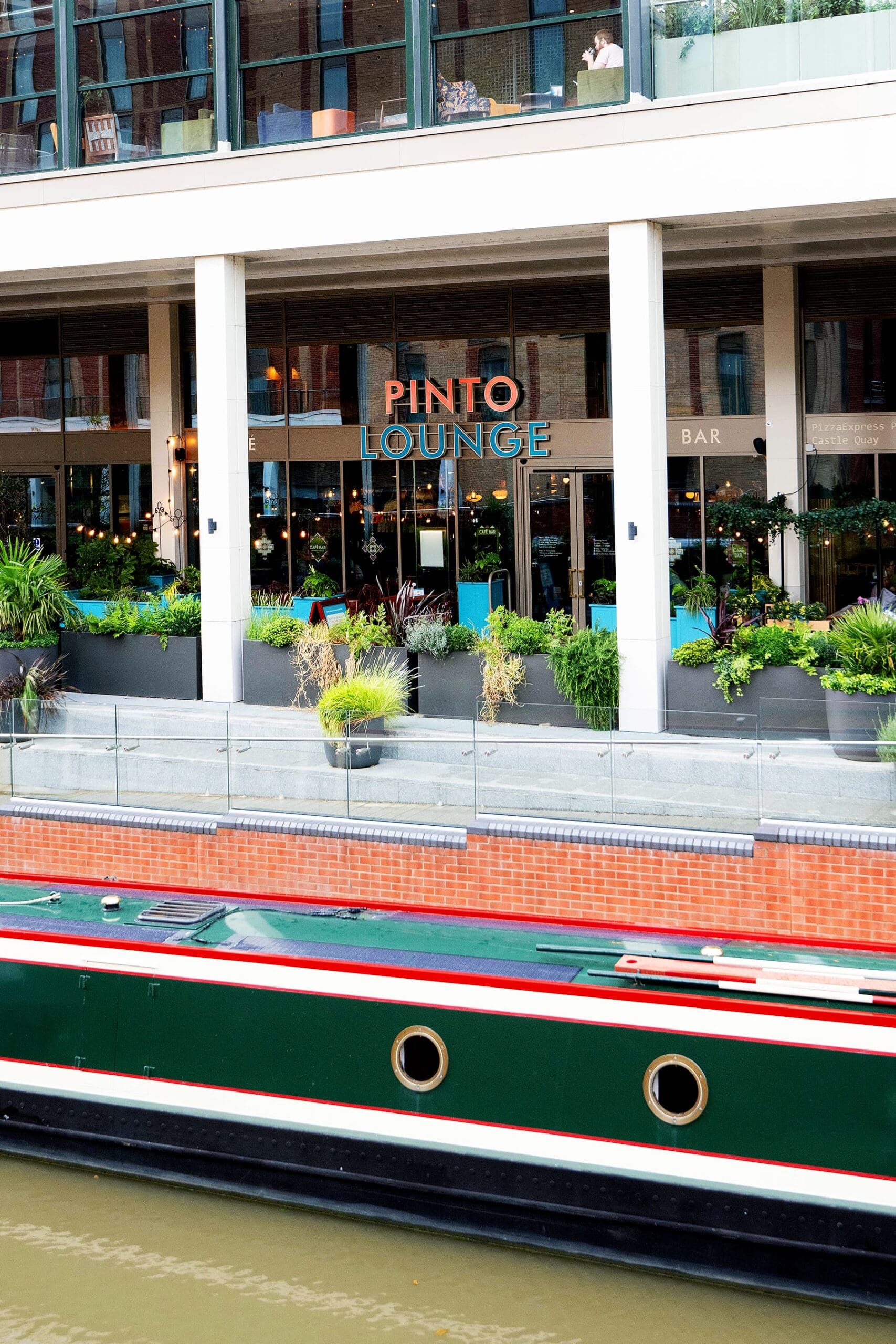 A canal boat floats on the water in front of Pinto Lounge, a modern café-bar with large windows, lush green plants, and outdoor seating. The Pinto building features a balcony with people visible inside.