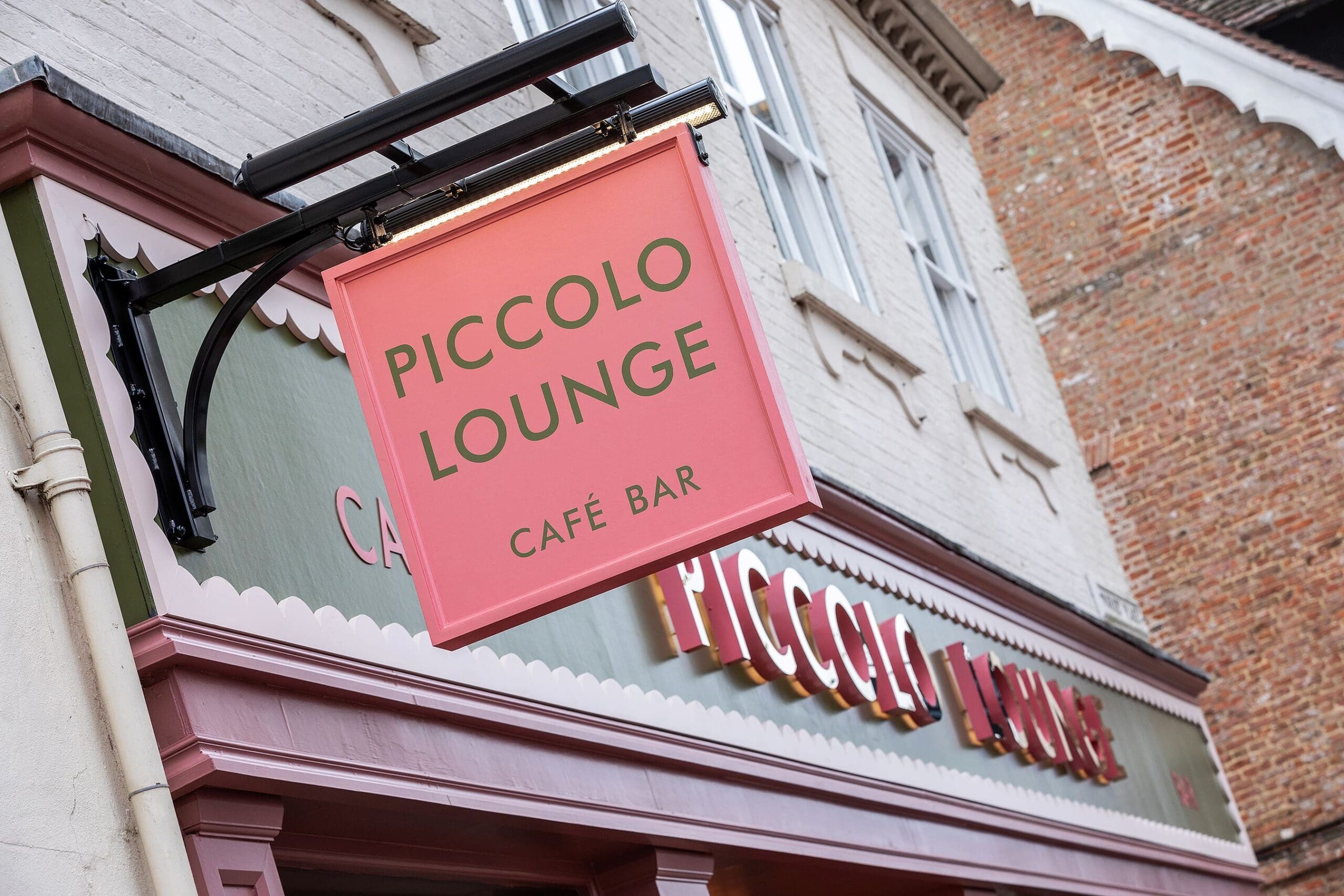 A pink hanging sign reading "Piccolo Lounge Café Bar" is displayed outside a café, with the same piccolo lounge branding visible on the storefront below. The building features white and brick walls.