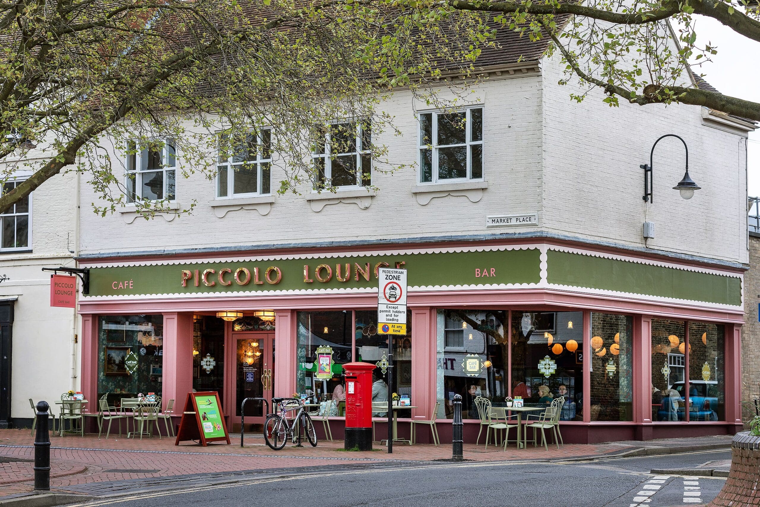 Street view of Piccolo Lounge café and bar on a corner, with outdoor tables and chairs, a red post box, a bicycle, and leafy trees overhead. The white building with green and pink trim highlights the inviting vibe of Piccolo Lounge.