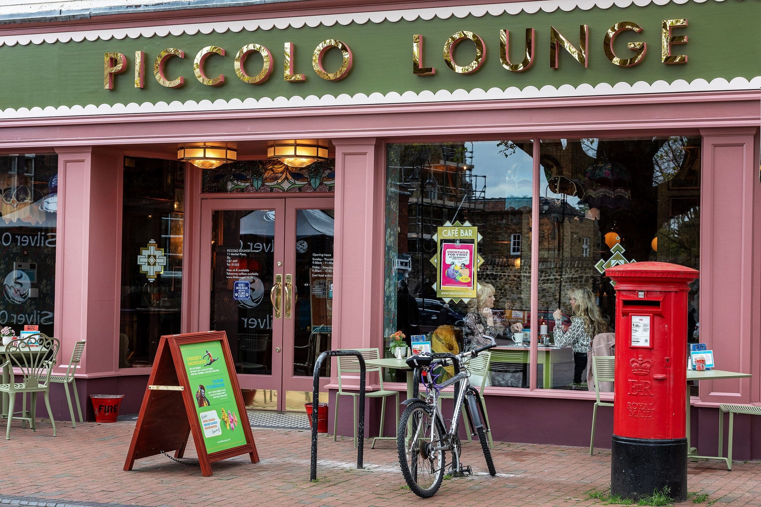 Street view of Piccolo Lounge café with large glass windows, outdoor seating, a classic red postbox, a bicycle locked to a bike rack, and a green sandwich board sign on the brick pavement outside Piccolo Lounge.