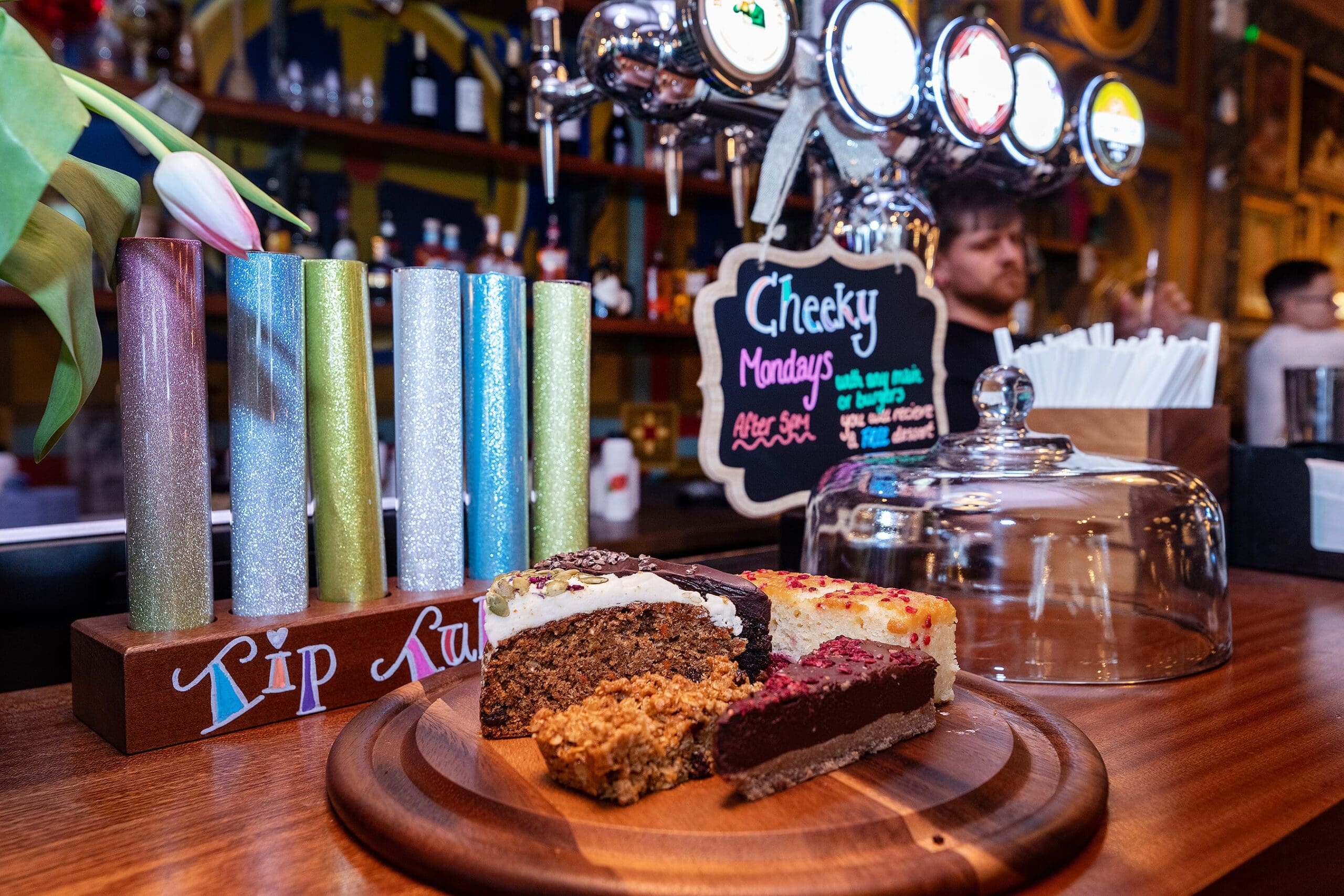 A wooden counter at Piccolo Lounge displays four assorted cake slices on a round board, next to glittery candles, a glass dome, and a sign reading "Cheeky Mondays." Bar taps and blurred patrons appear in the background.