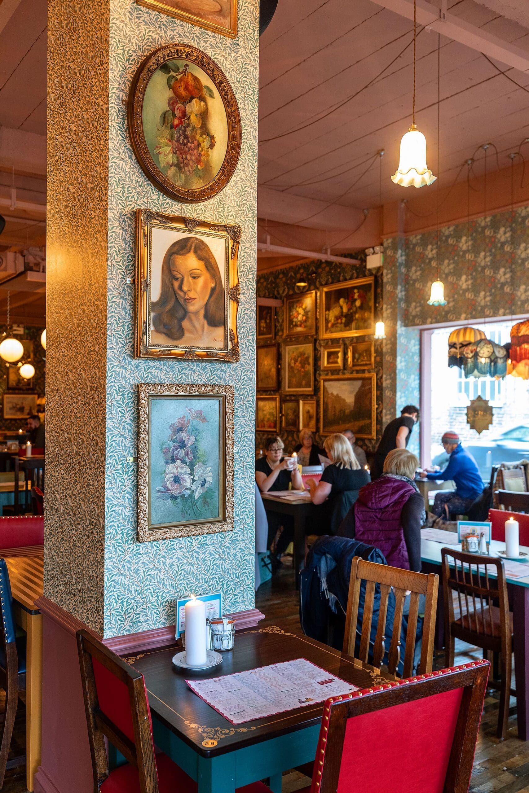 A cozy café interior with vintage decor, floral wallpaper, and framed art on the walls evokes a piccolo lounge vibe. A pillar features portraits and a flower painting above an empty table with red chairs and a menu.