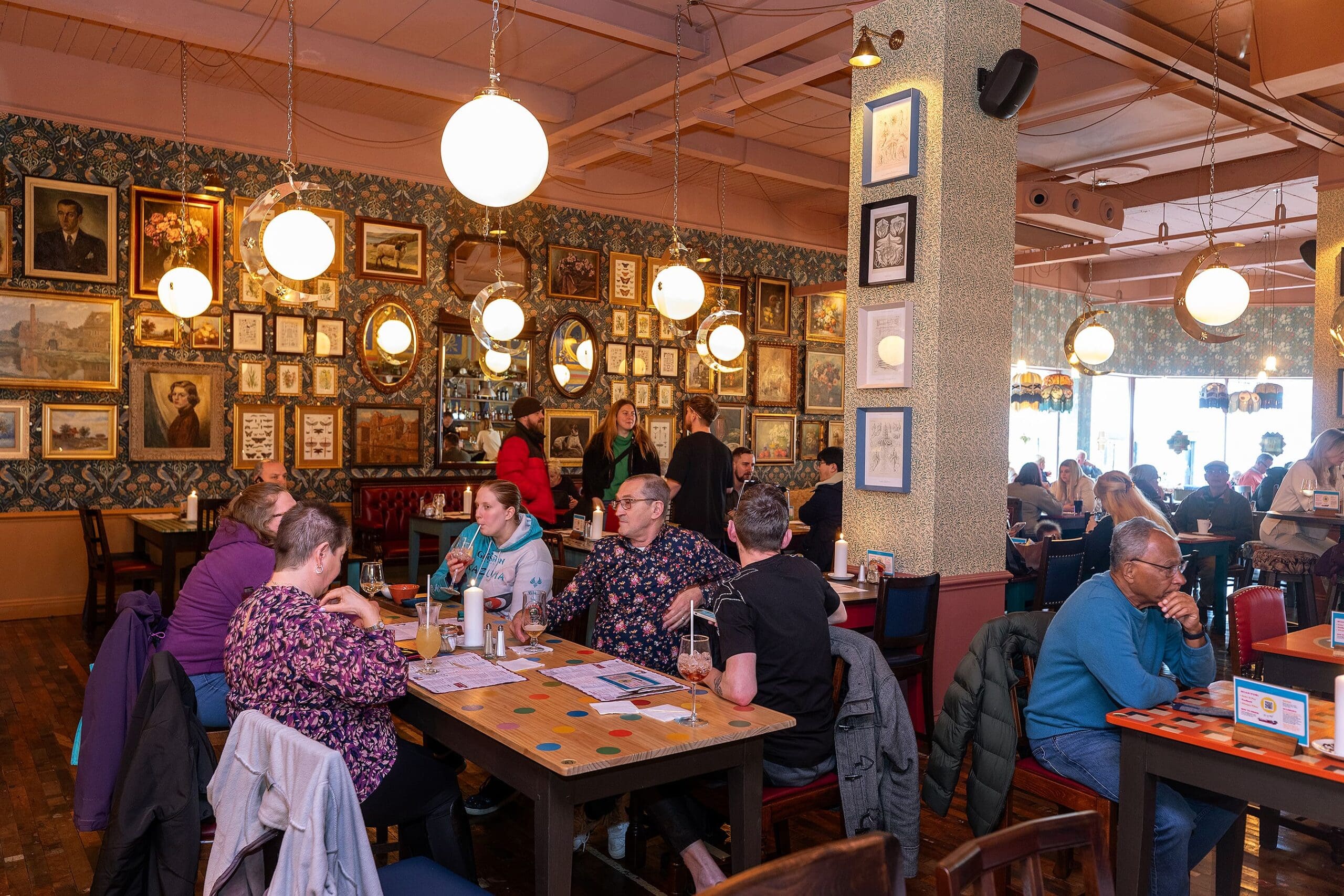 A lively café, reminiscent of a piccolo lounge, with groups of people at wooden tables, engaged in conversation. Framed pictures and portraits adorn the walls, while round pendant lights create a warm, inviting atmosphere.