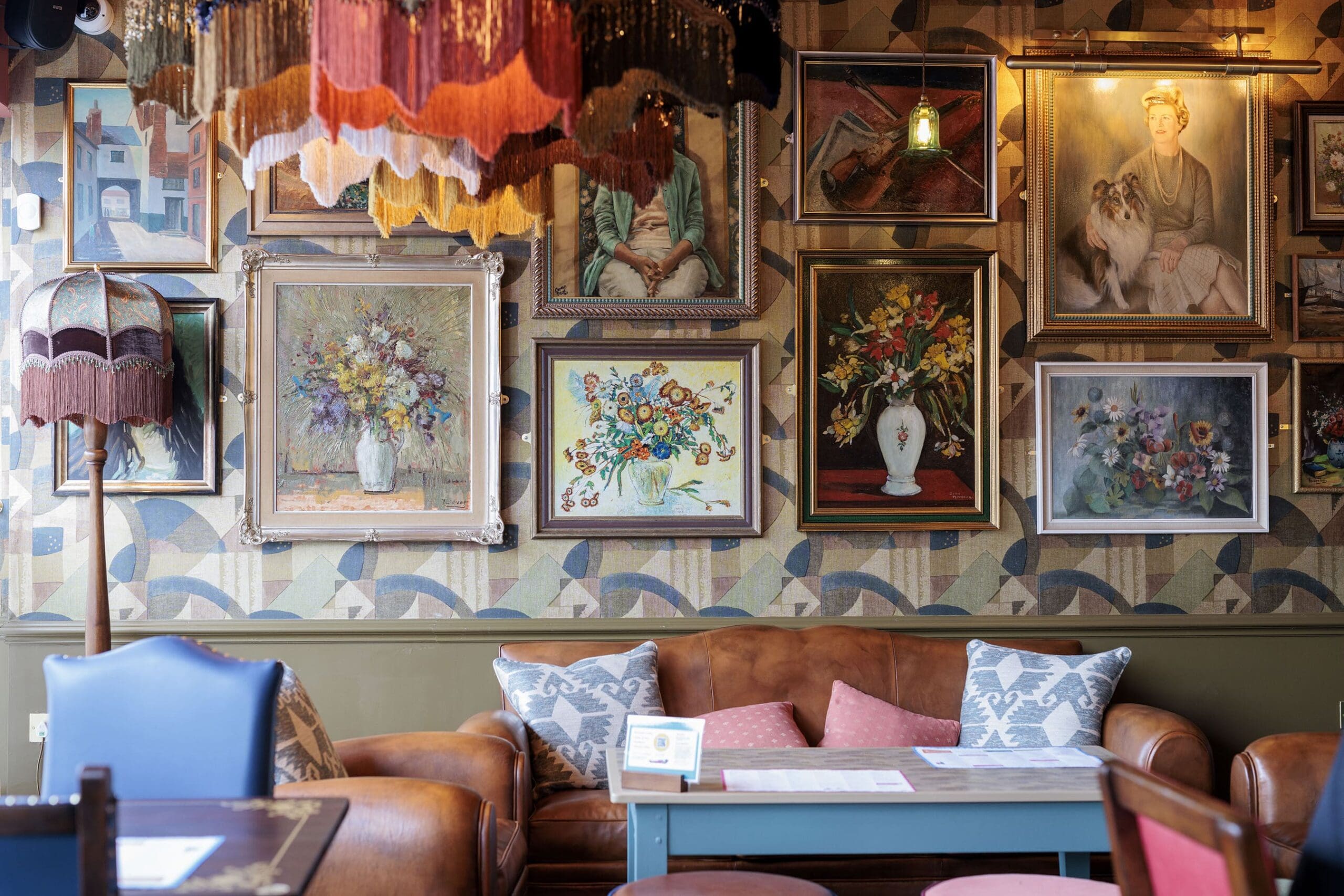Cozy cafe interior with a brown leather sofa, pastel cushions, a turquoise sarto table, and walls decorated with framed floral and portrait paintings. Vintage lamps with fringed shades hang from the ceiling and stand in the corner.