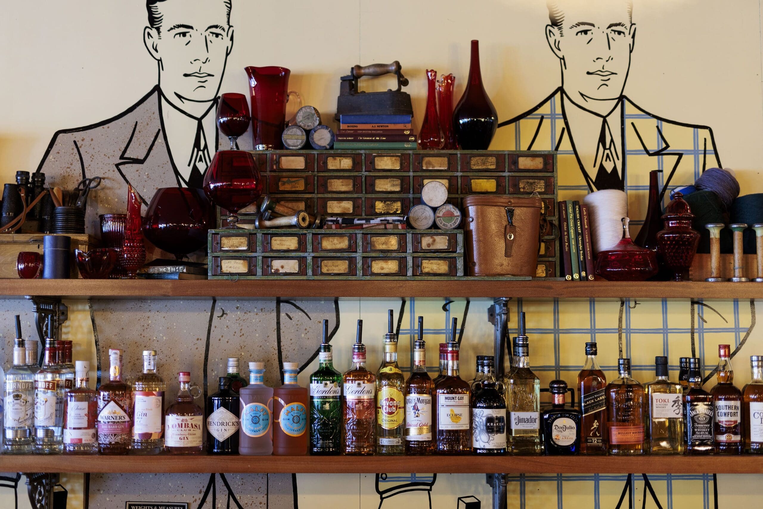 A shelf displays various liquor bottles, vintage apothecary drawers, red glassware, gauges, and decorative items against a wall featuring sarto-inspired line art of men in suits.