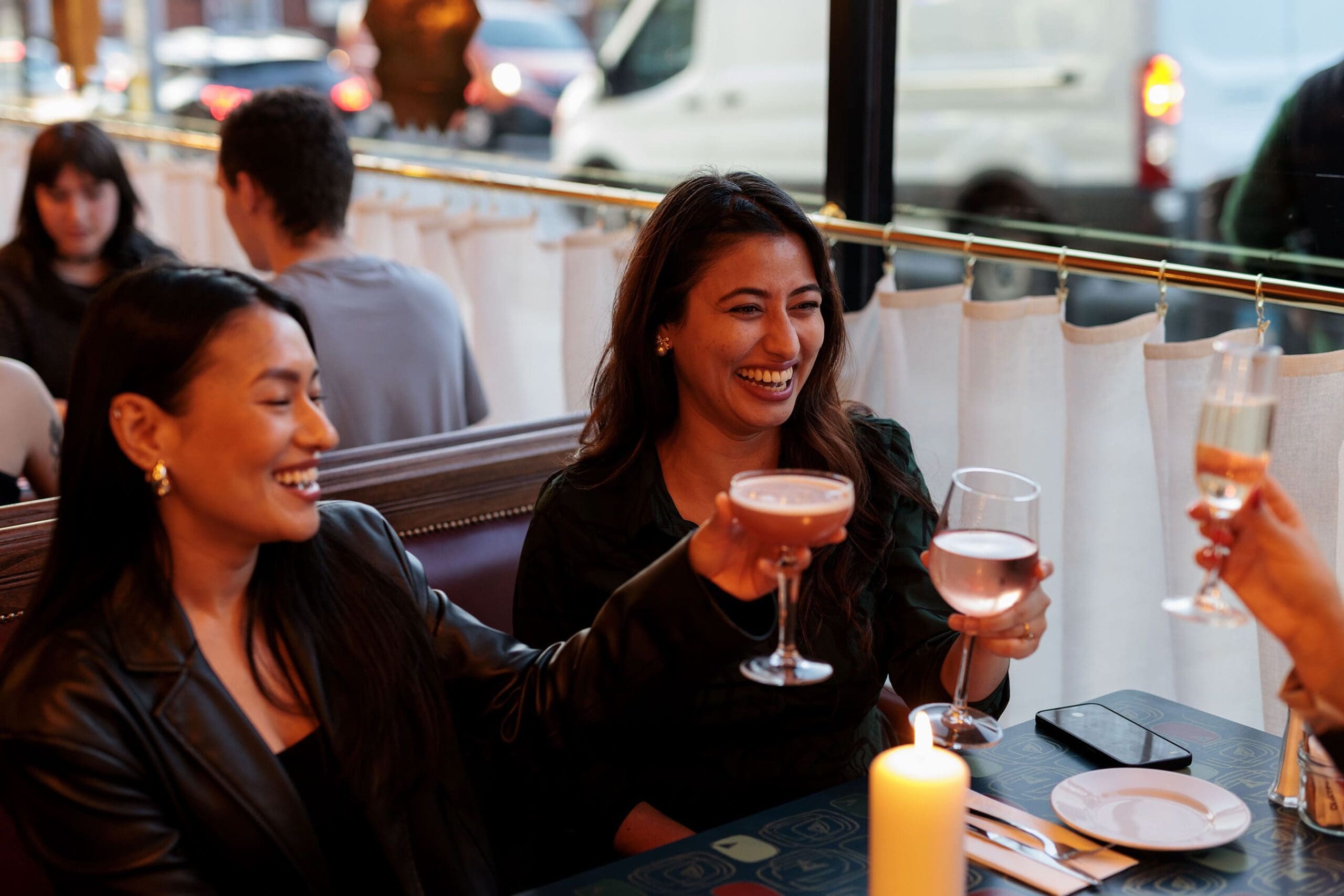 Two women sit at a sarto restaurant table, smiling and raising glasses in a lively toast with another guest. The warm setting, complete with candlelight and blurred diners in the background, creates a welcoming atmosphere.