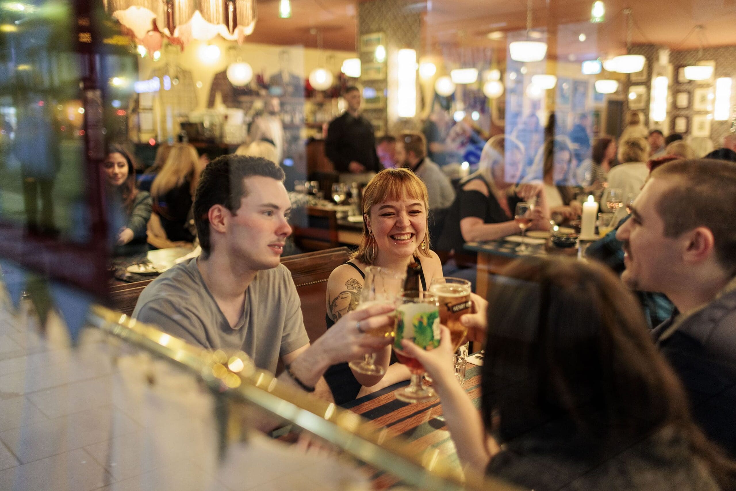Four friends sit at a Sarto restaurant table, smiling and raising their drinks in a toast. The atmosphere is lively and warm, with other diners and cozy lighting in the background.
