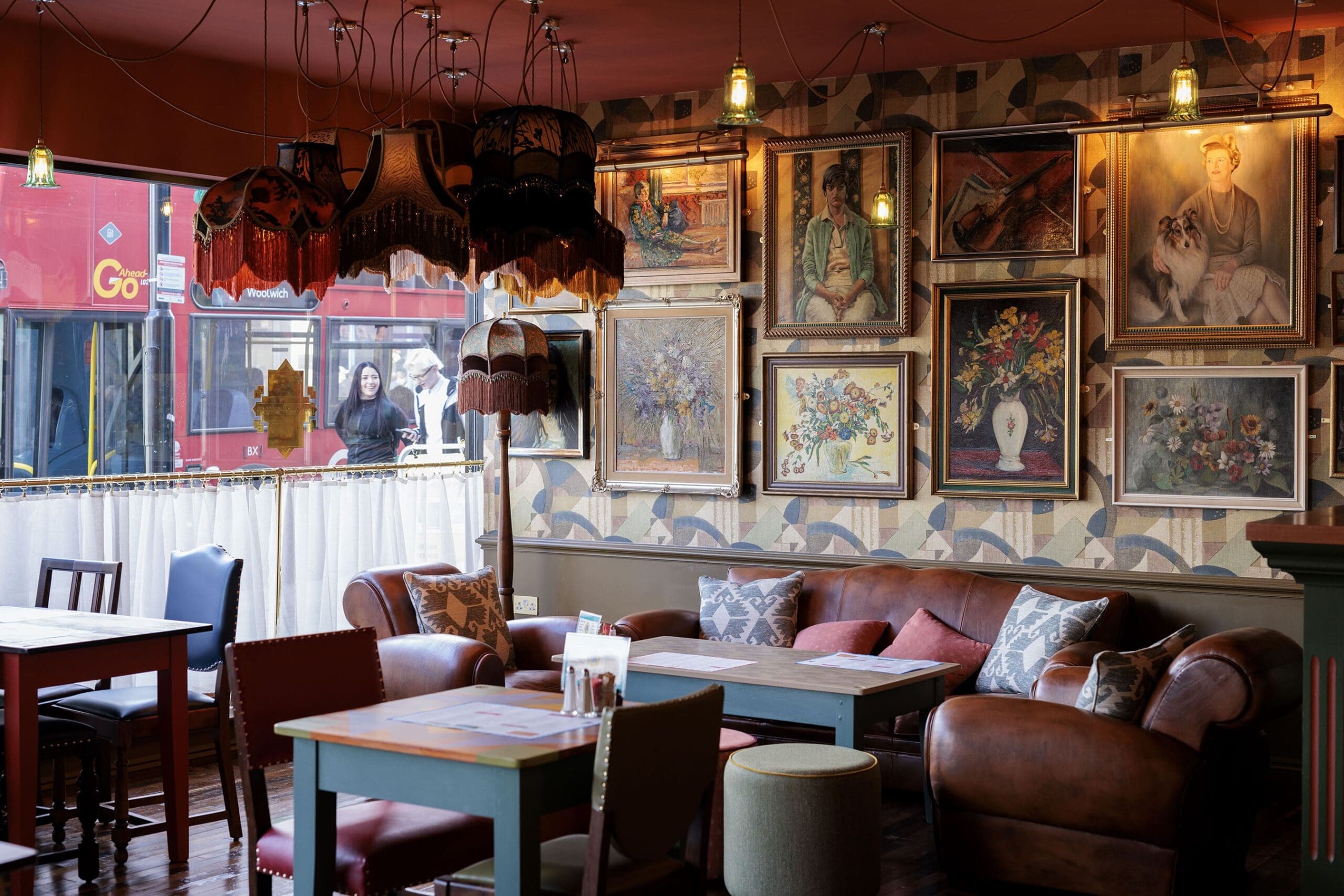 A cozy sarto cafe interior with leather sofas, wooden tables, eclectic lamps, and a gallery wall of paintings. Large windows reveal a red London bus and people passing by outside. Warm, inviting, and vintage atmosphere.