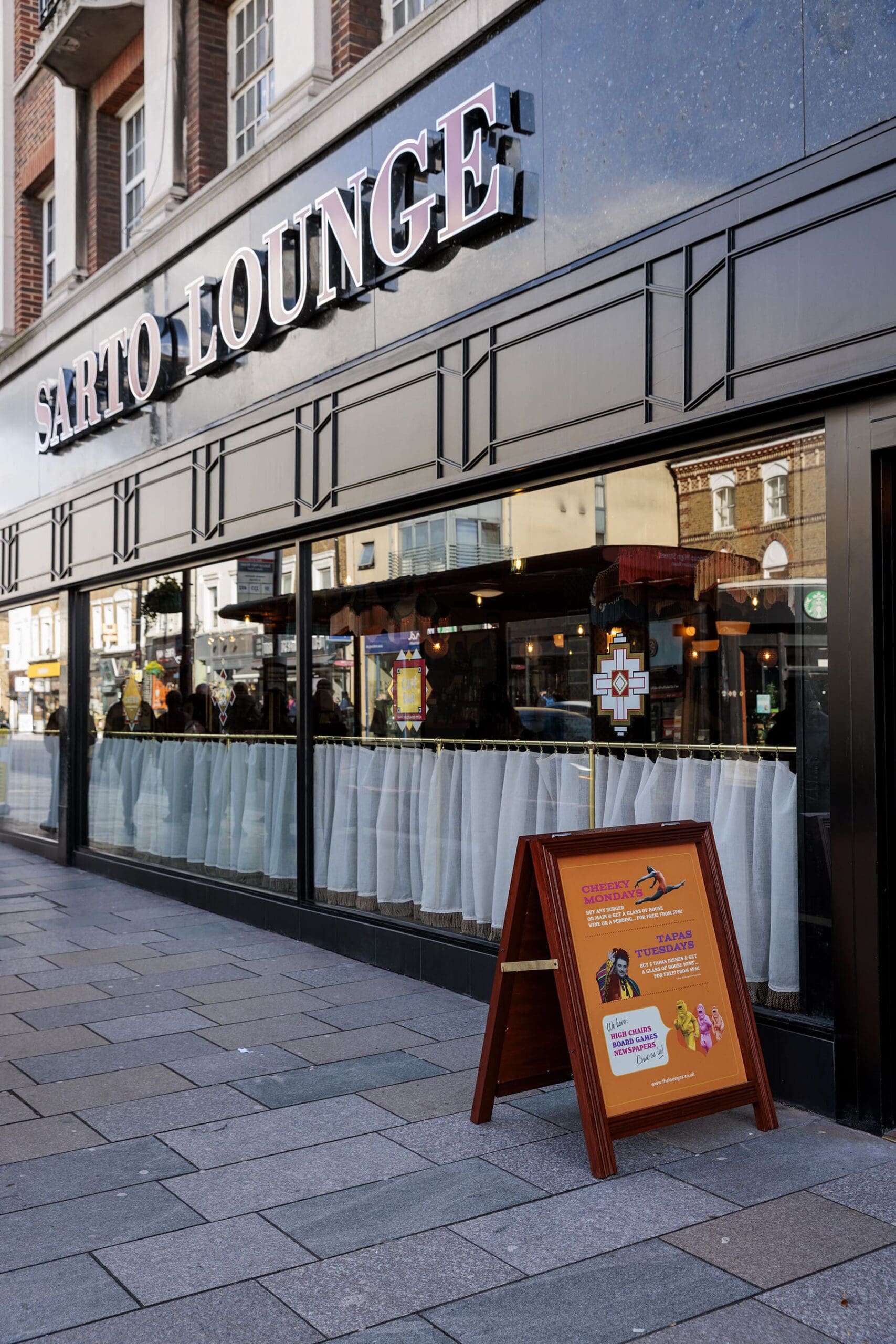 A street view of Sarto Lounge café with large windows, white curtains, and a sidewalk sign advertising Sarto’s food and events standing outside the entrance.