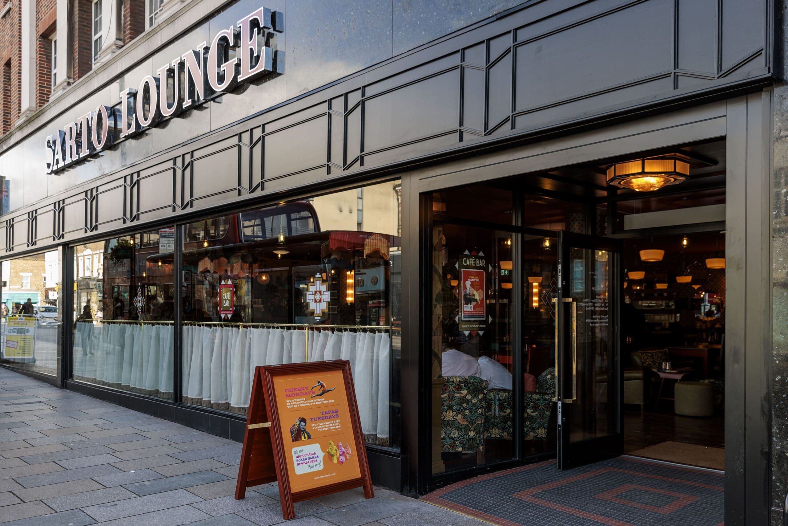 Street view of the Sarto Lounge café with large glass windows, a visible interior lit warmly, and a sandwich board outside on the sidewalk advertising community events at Sarto.