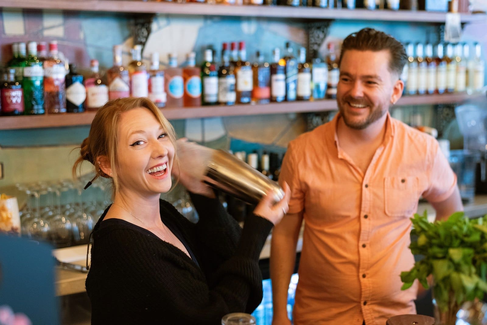 A smiling woman shakes a cocktail shaker behind a bar, while a man in a peach shirt stands beside her, smiling. Bottles and glasses line the shelves in the background, including an elegant Barolo among the selection.