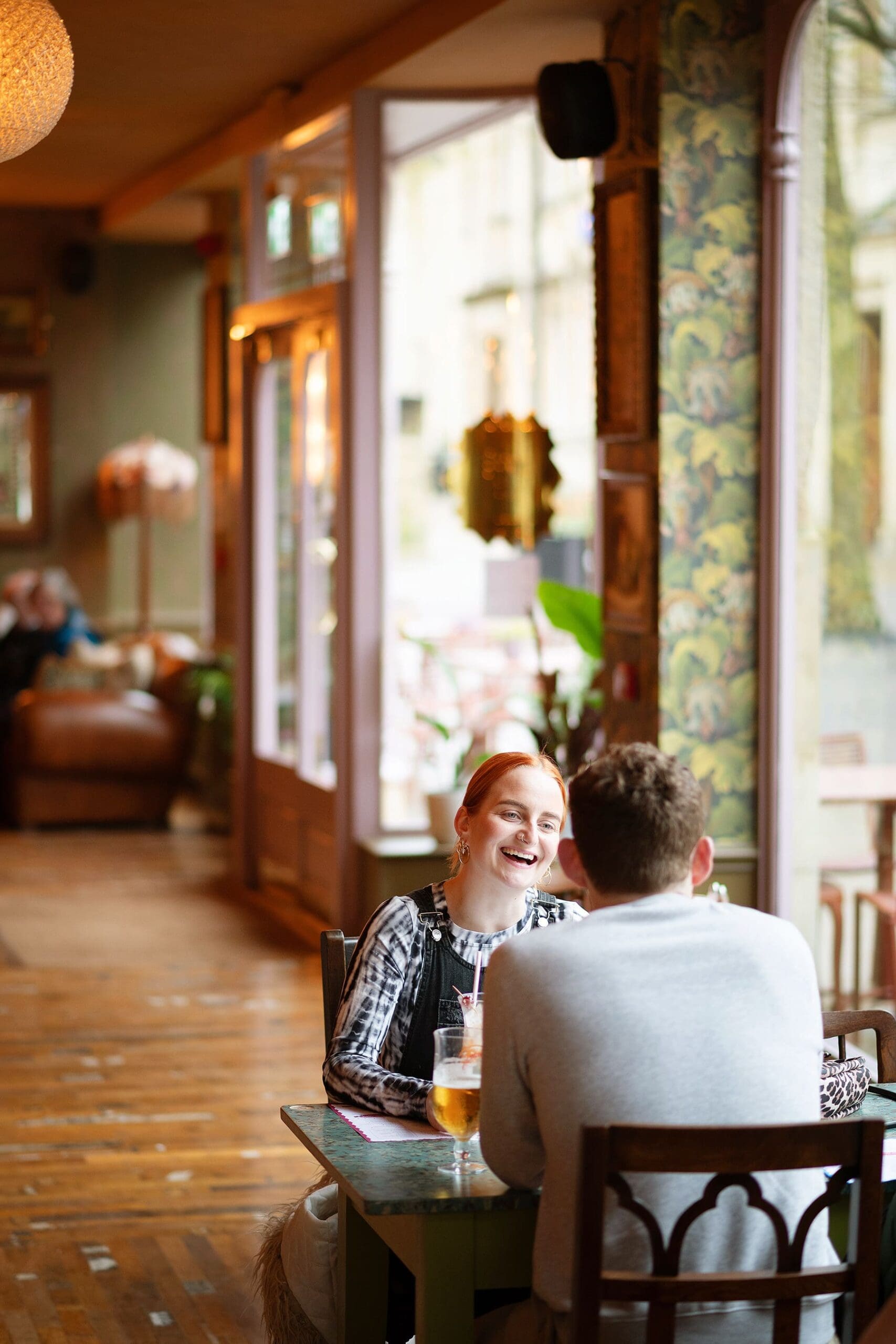 A young woman and man sit across from each other at a small table in a cozy cafe, smiling and talking, with drinks in front of them. The scene captures the warmth and connection at the heart of our About Us story.