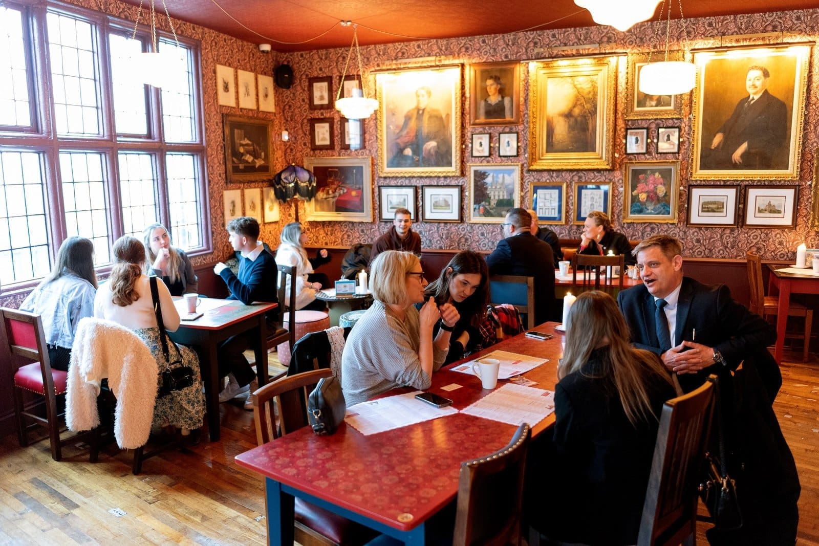 People sit at tables in a warmly lit café or restaurant with red walls decorated with framed portraits and artwork, sipping Barolo and engaging in lively conversations. Large windows let in natural light.