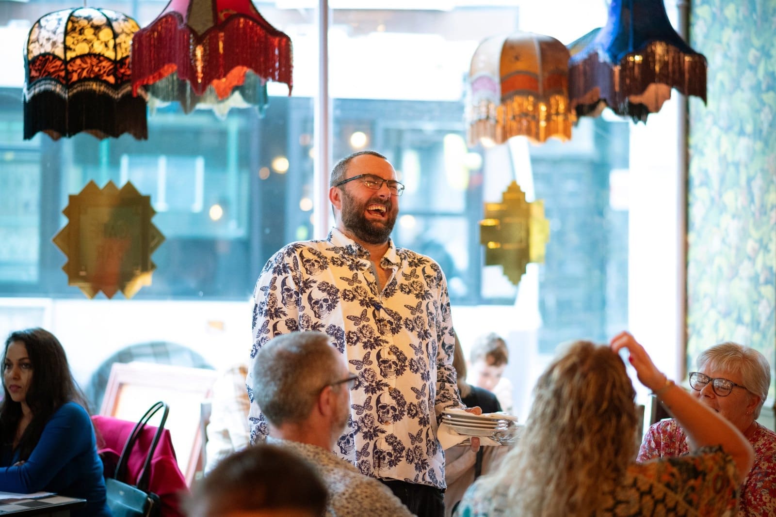 A man in a patterned shirt laughs while standing in a lively, colorful cafe with vintage lamps and people seated at tables around him, savoring a glass of Barolo.