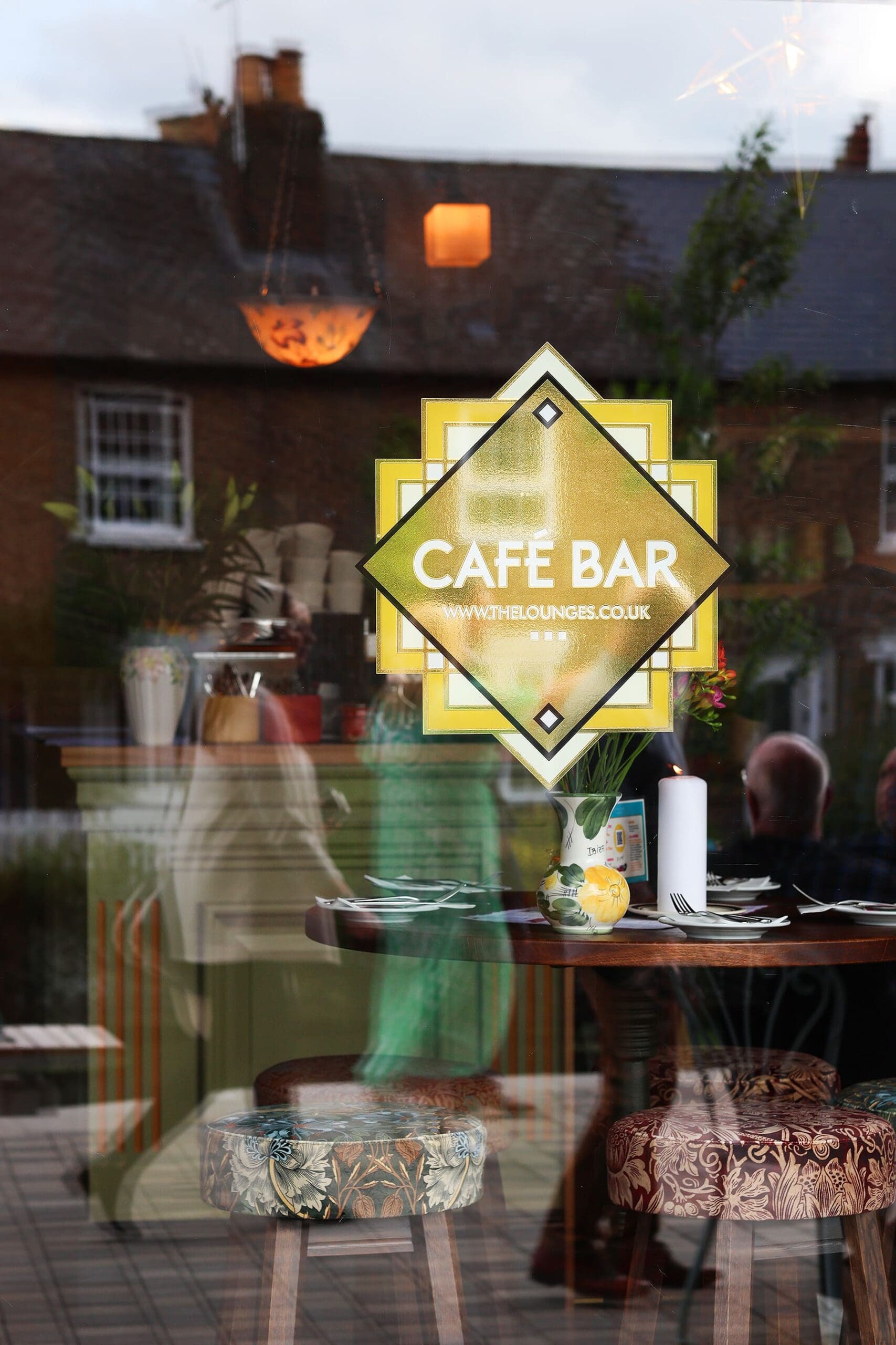 A glass window displays a yellow "Café Bar" sign. Inside, a table is set with plates and cutlery, surrounded by patterned Quattro stools. Outside, houses and trees are reflected in the glass.