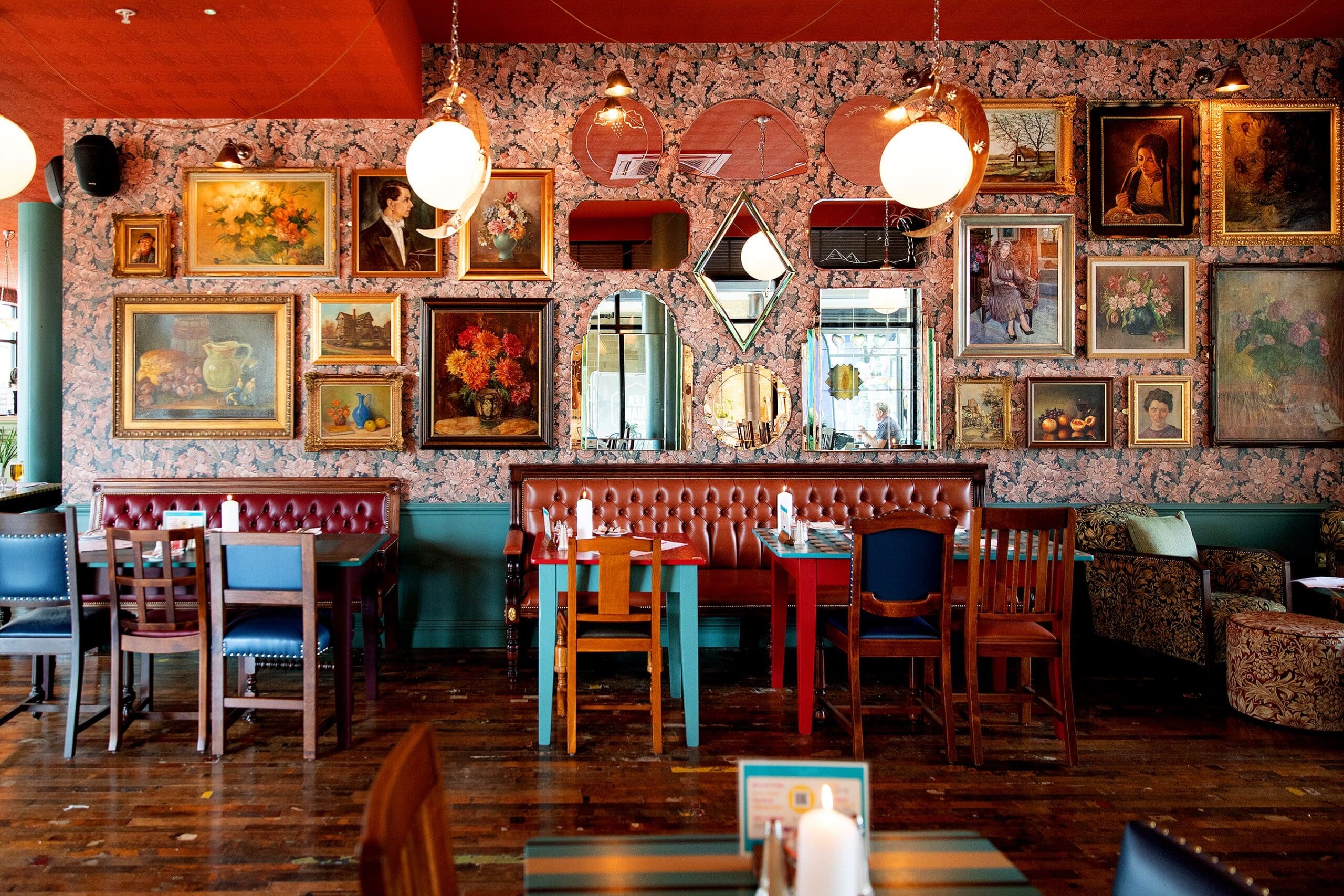 A cozy Quattro restaurant with eclectic decor, featuring mismatched chairs, a brown tufted bench, and walls covered in framed art and mirrors. The space is warmly lit by hanging globe lamps.