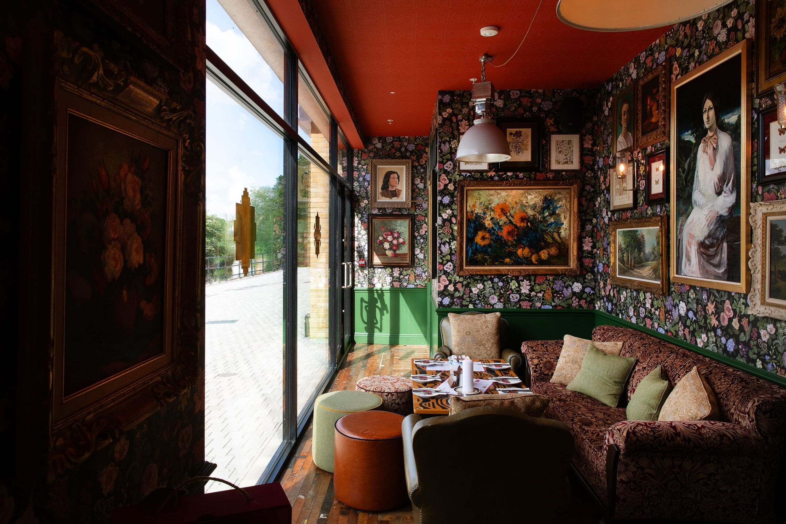 A cozy, vintage-style café with floral wallpaper, eclectic framed art, a plush Quattro sofa, pillows, and small tables by large floor-to-ceiling windows letting in natural light.