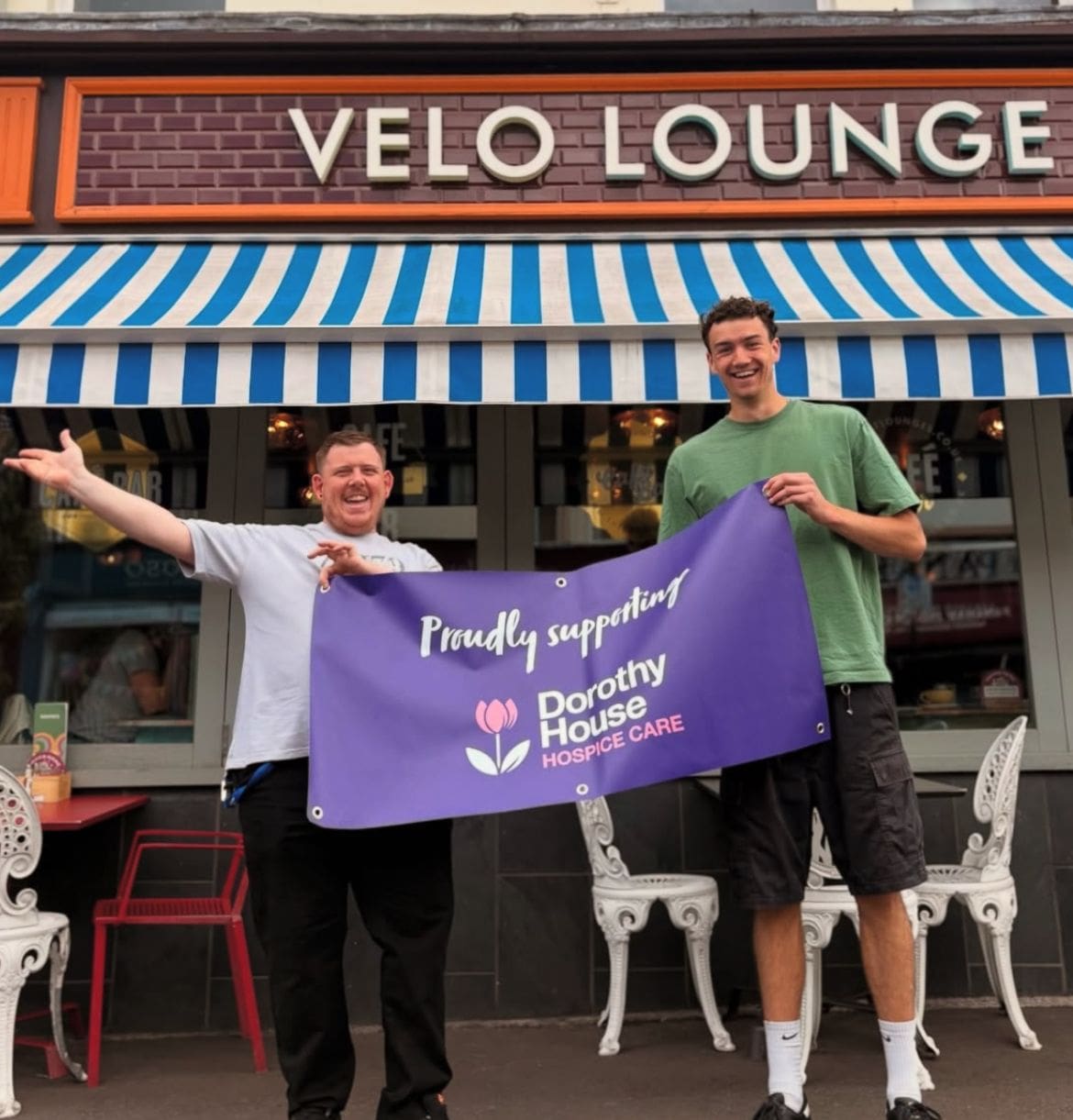 Two smiling men stand outside Velo Lounge, holding a purple banner that reads "Proudly supporting Dorothy House Hospice Care" with a tulip logo. Blue and white striped awning and outdoor seating are visible behind them.