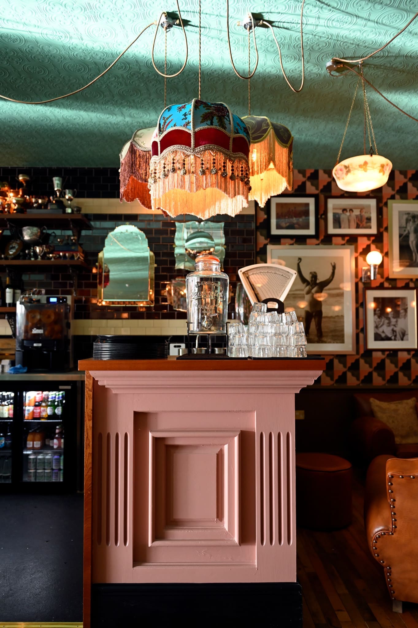 A vintage-style café interior with a pink counter, Verdetto stacked glasses, a blender, and an ornate fringed lampshade. The walls display framed pictures, mirrors, and dark tiles beneath a patterned teal ceiling.