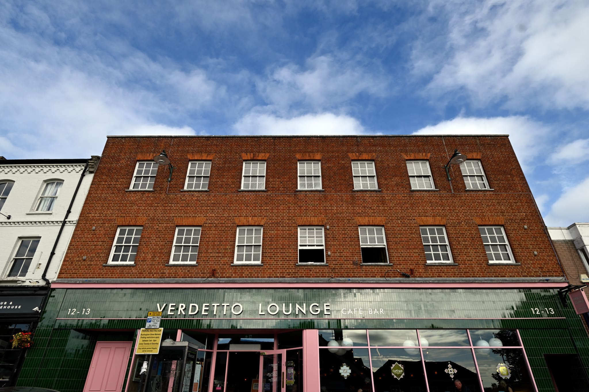 A three-story brick building with green tiles and large windows houses "Verdetto Lounge Café Bar." The sky above is partly cloudy, and the Verdetto building is flanked by neighboring shops.