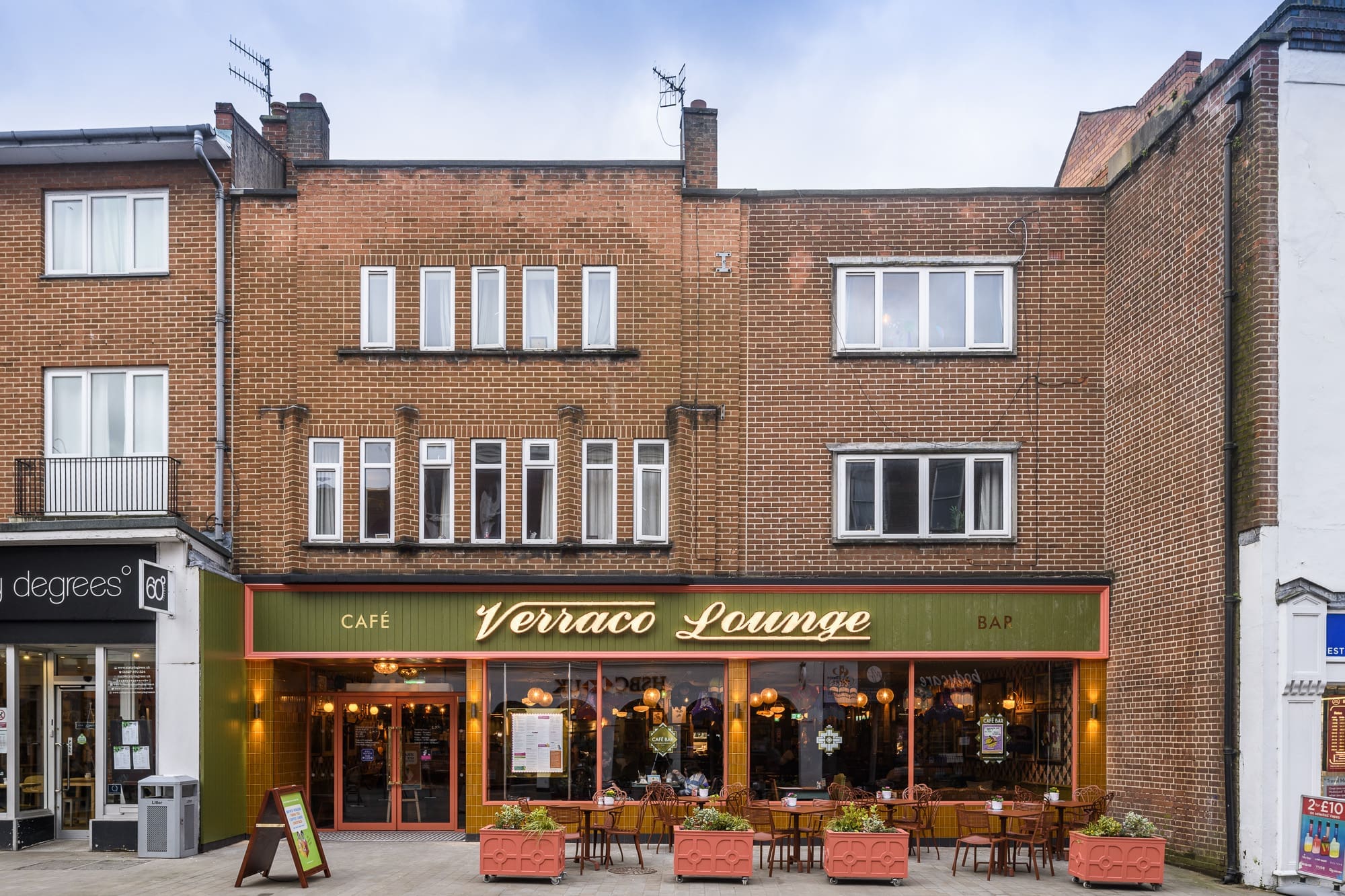 Street view of Verraco Lounge, a café and bar with large windows, verraco-red planters framing outdoor seating, and warm lighting inside. The brick building features apartments above the inviting street-level businesses.