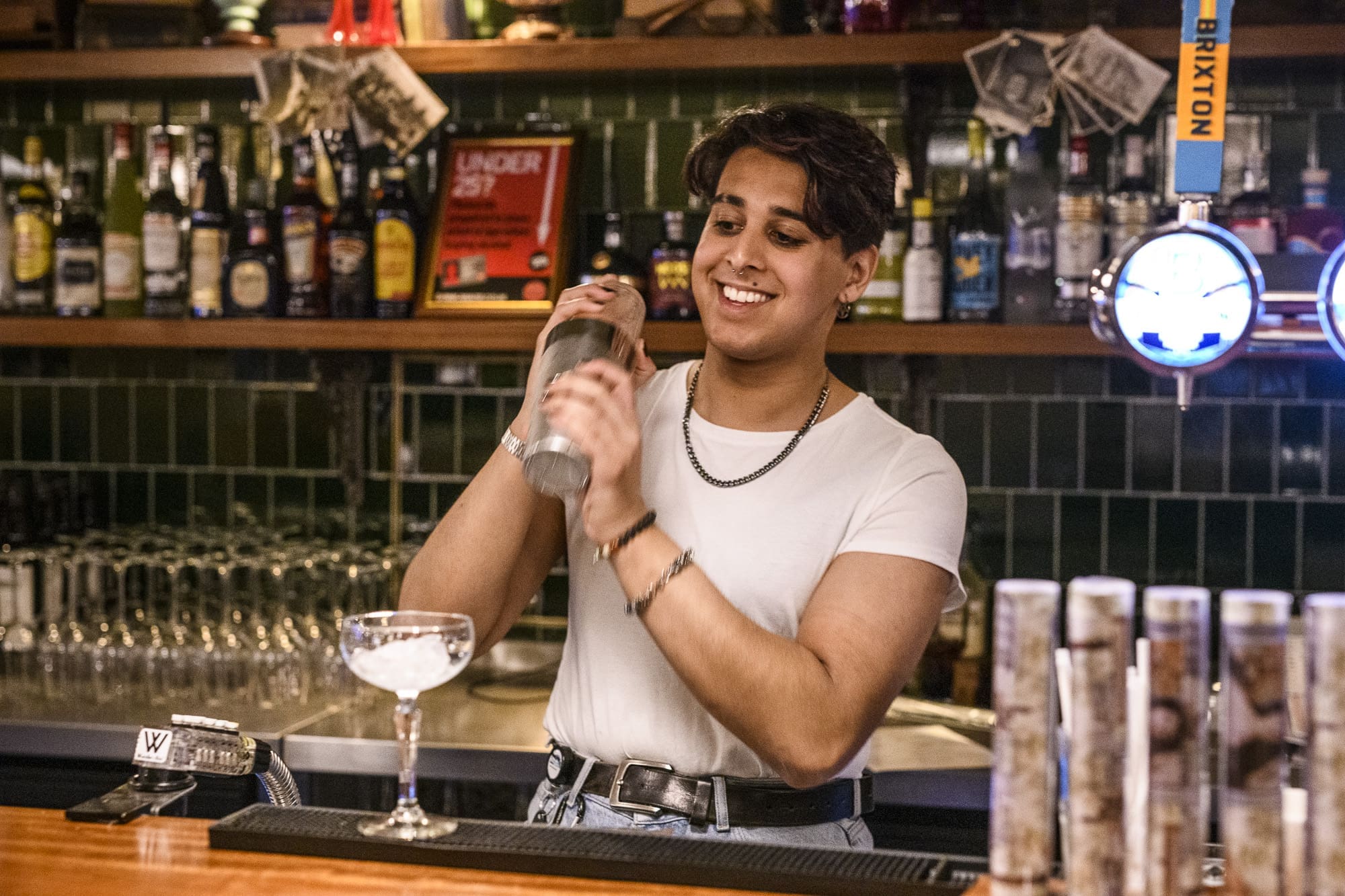 A smiling bartender in a white t-shirt shakes a verraco-branded cocktail shaker behind the bar, preparing a drink. Shelves of bottles and glasses are visible in the background.