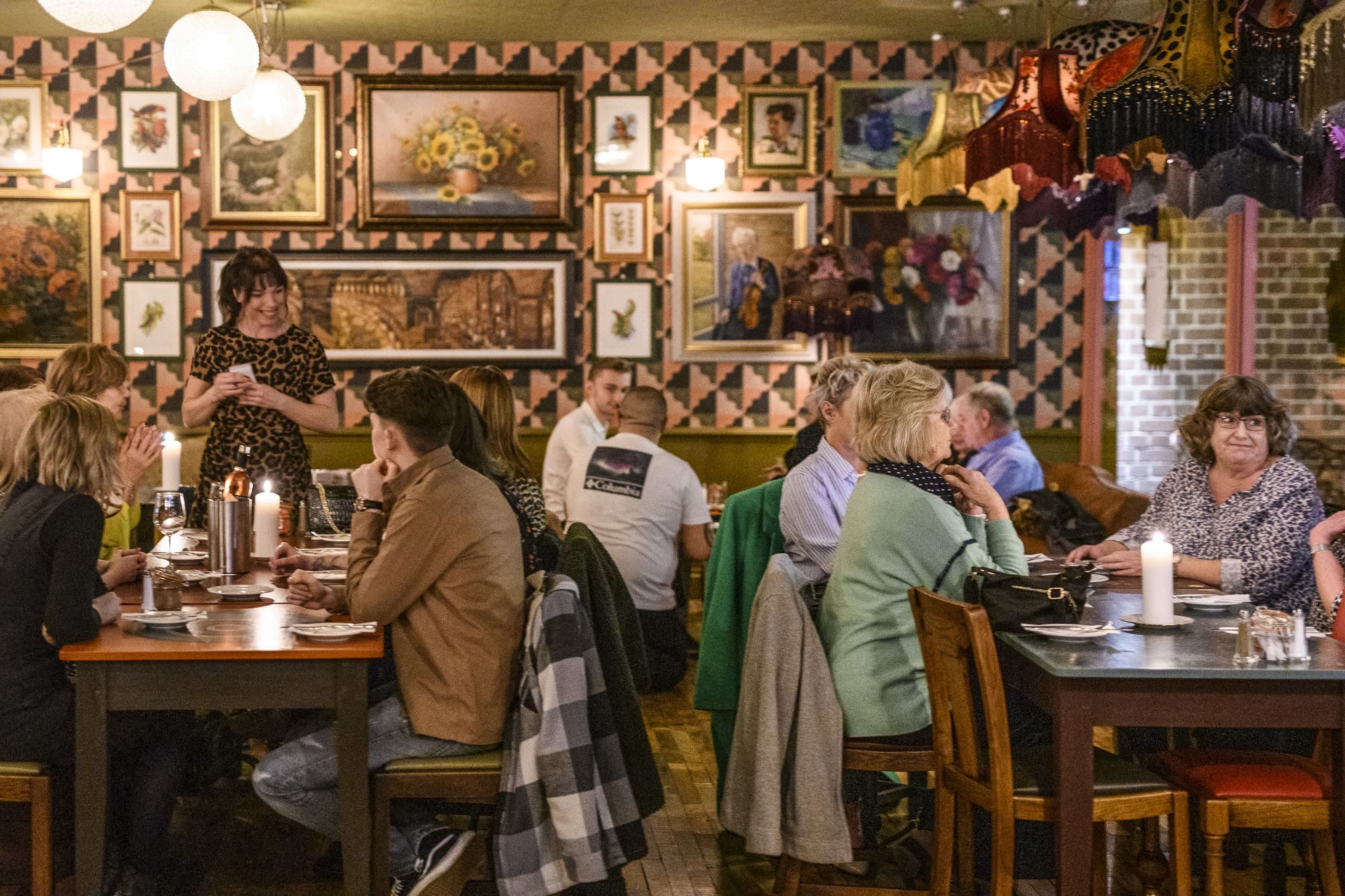 A lively restaurant with groups of people dining and talking at tables, surrounded by eclectic wall art, patterned wallpaper, and hanging lampshades. At the center, a verraco statue adds character as a waitress engages with guests.