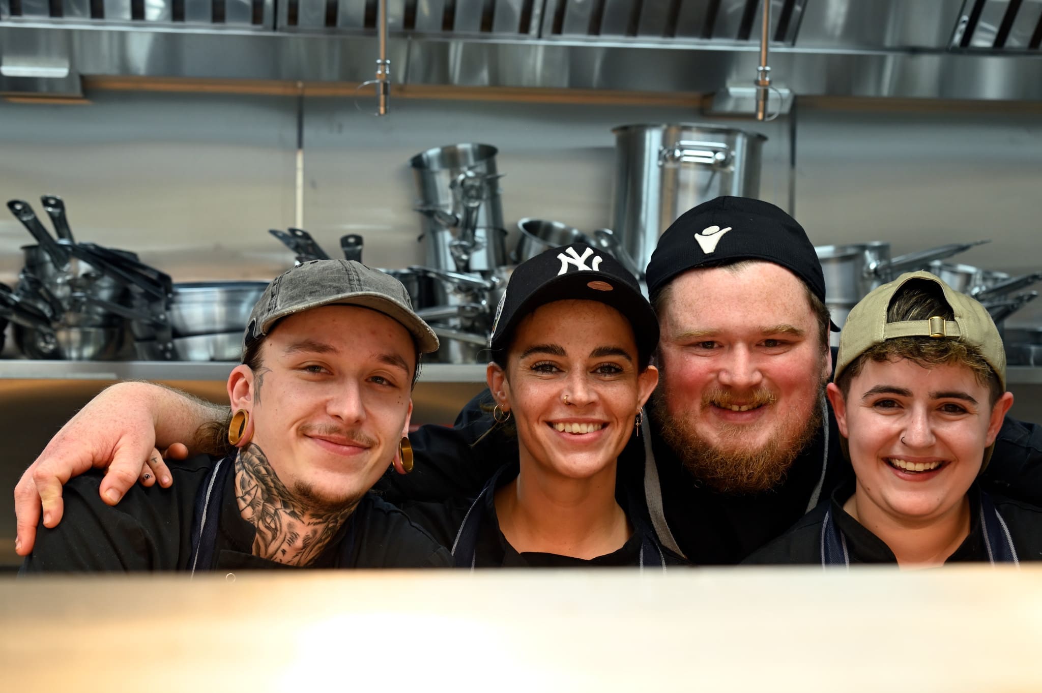 Four smiling chefs stand close together in the Viento Lounge kitchen, arms around each other, with metal pots and pans hanging in the background. They wear aprons, caps, and look happy and friendly.