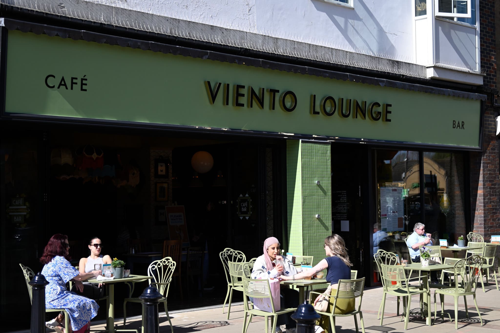 People sit at green outdoor tables outside Viento Lounge on a sunny day, chatting and enjoying drinks. The café bar’s green sign and large windows stand out as sunlight casts shadows on the pavement.