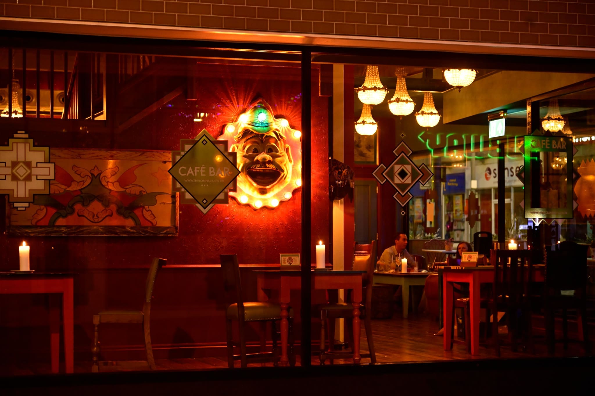 Warmly lit Vintro café interior seen through a window at night, featuring a glowing mask wall decoration, candles on tables, chandeliers, and a few people seated inside.