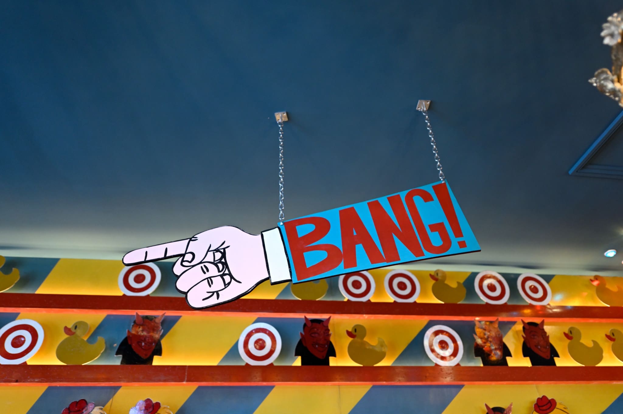 A colorful Vintro sign with a cartoon hand pointing and the word “BANG!” in bold red letters hangs from the ceiling; behind it are rows of yellow duck targets and red animal heads on a carnival-style wall.