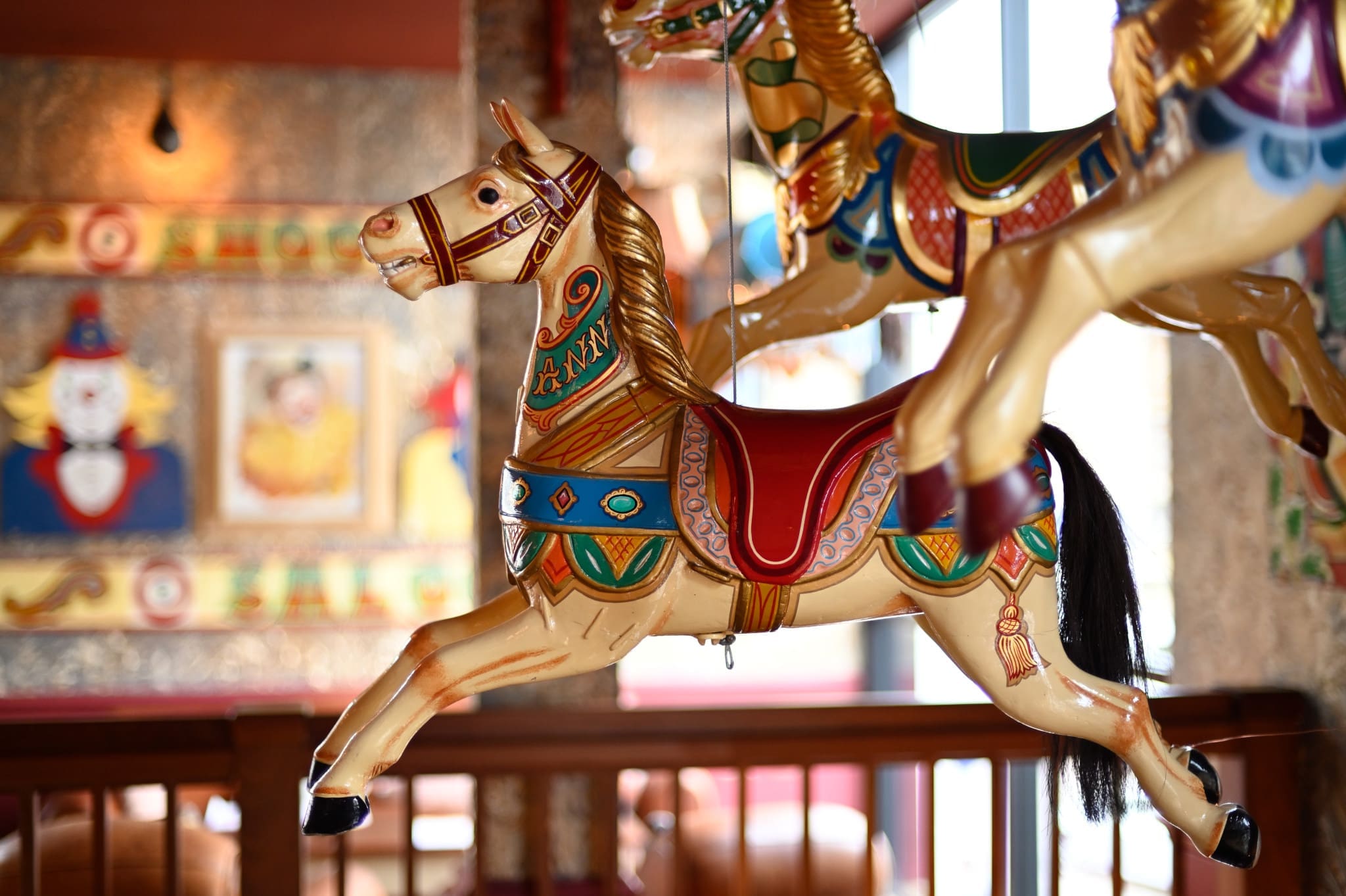 A close-up of colorful Vintro carousel horse figures indoors, featuring ornate patterns and a blurred background with circus-themed wall art and warm lighting.
