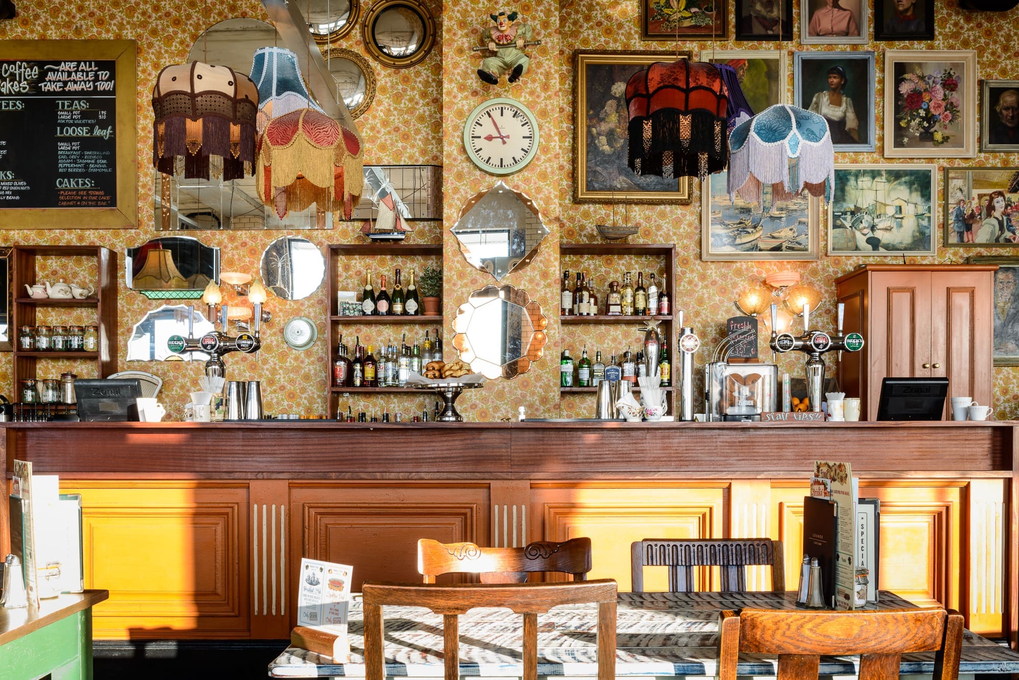 A cozy vintage café interior with patterned wallpaper, eclectic lampshades, and framed paintings creates a charming Visto. Sunlight streams in past mirrors behind a wooden bar stocked with bottles, illuminating tables and chairs in the foreground.