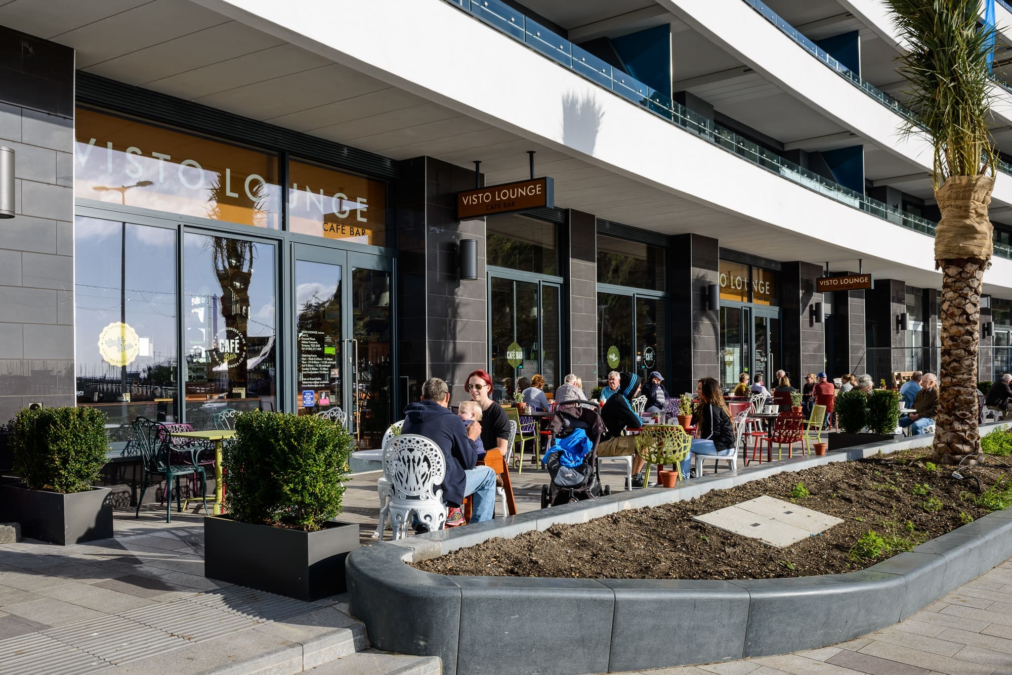 People relax at Visto Lounge’s outdoor tables, savoring food and drinks on a sunny day. The café, with its large windows and potted plants, is set in a modern building with balconies above.