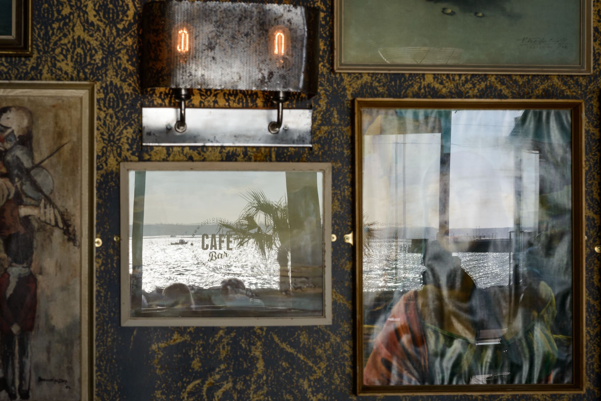Framed pictures and a lamp on a patterned wall, with a window reflecting the sea, a palm tree, and parts of a café sign—Visto blends indoor décor with an inviting outdoor water view.