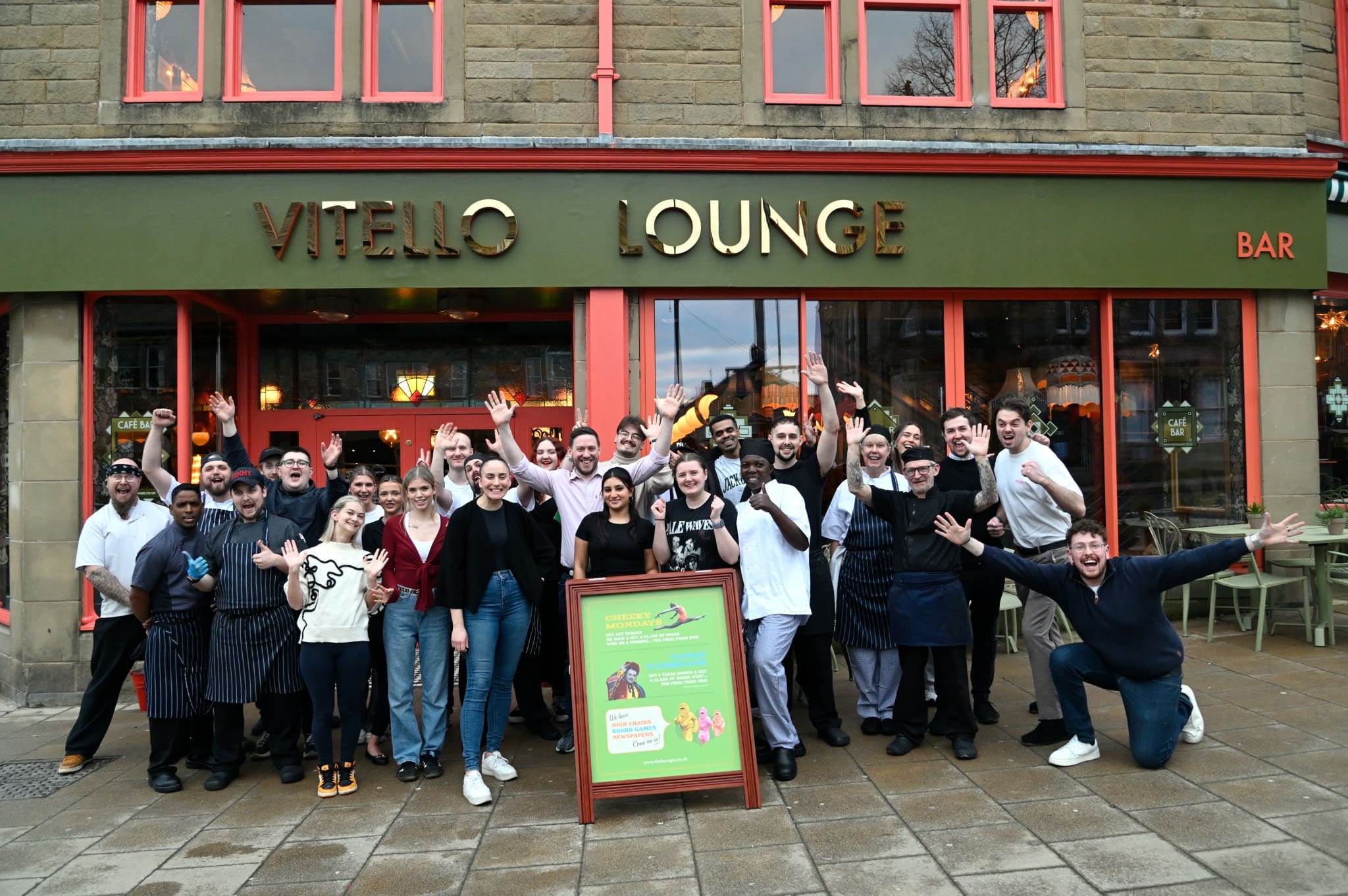 A large group of people, likely staff, stand smiling and waving in front of Vitello Lounge on a city street, with a sandwich board sign displayed in the center, highlighting the vibrant spirit of Vitello.