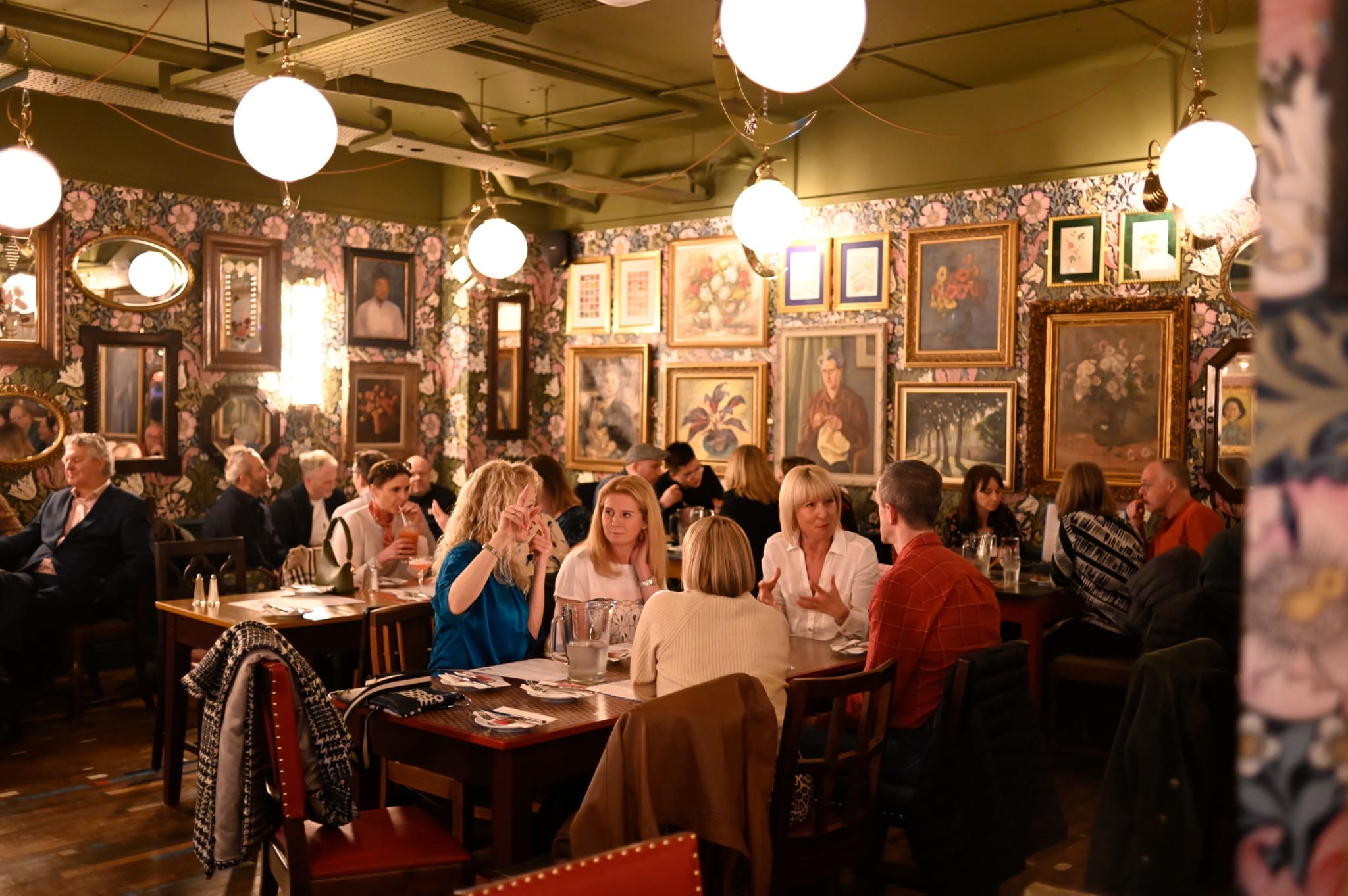 People are dining and conversing in a warmly lit, cozy restaurant with patterned wallpaper, walls adorned with framed paintings and mirrors, and tables featuring dishes like vitello. The atmosphere appears lively and social.