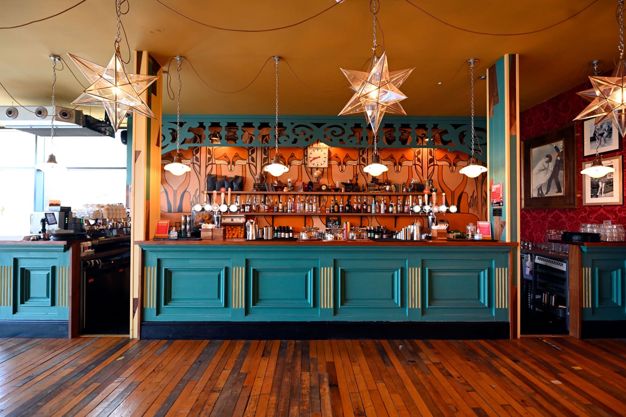 A stylish bar with teal panels, warm wood floors, star-shaped pendant lights, and shelves of bottles. Ornate decor and vintage touches create a cozy, eclectic Volpo vibe that's perfect for relaxing or socializing.