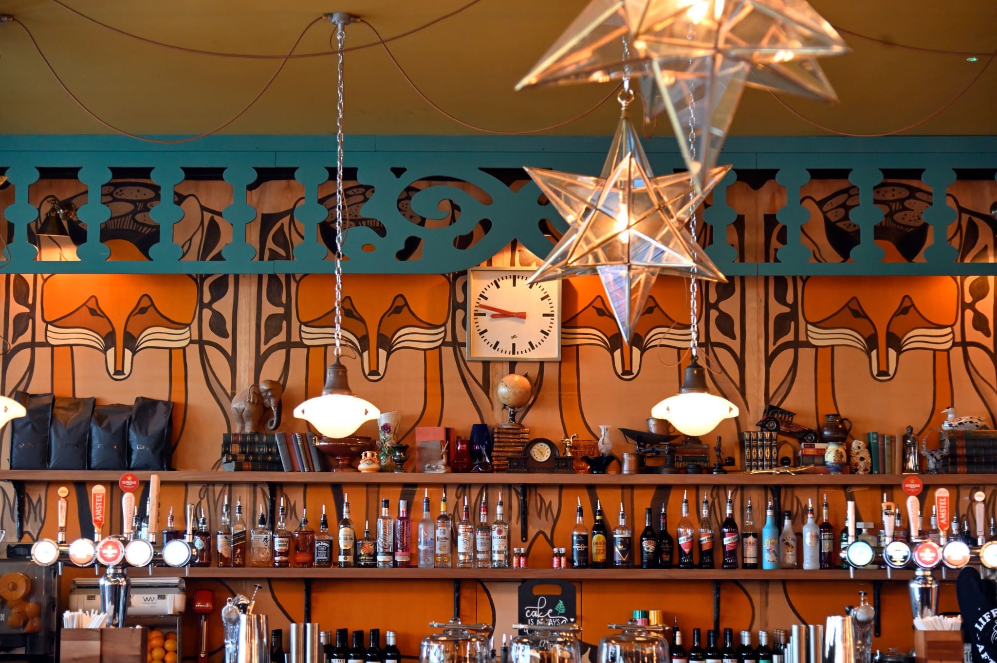 A vibrant Volpo bar interior with hanging star-shaped lights, a central clock, and colorful Art Nouveau-style decor. Shelves display various liquor bottles while beer taps line the counter below.