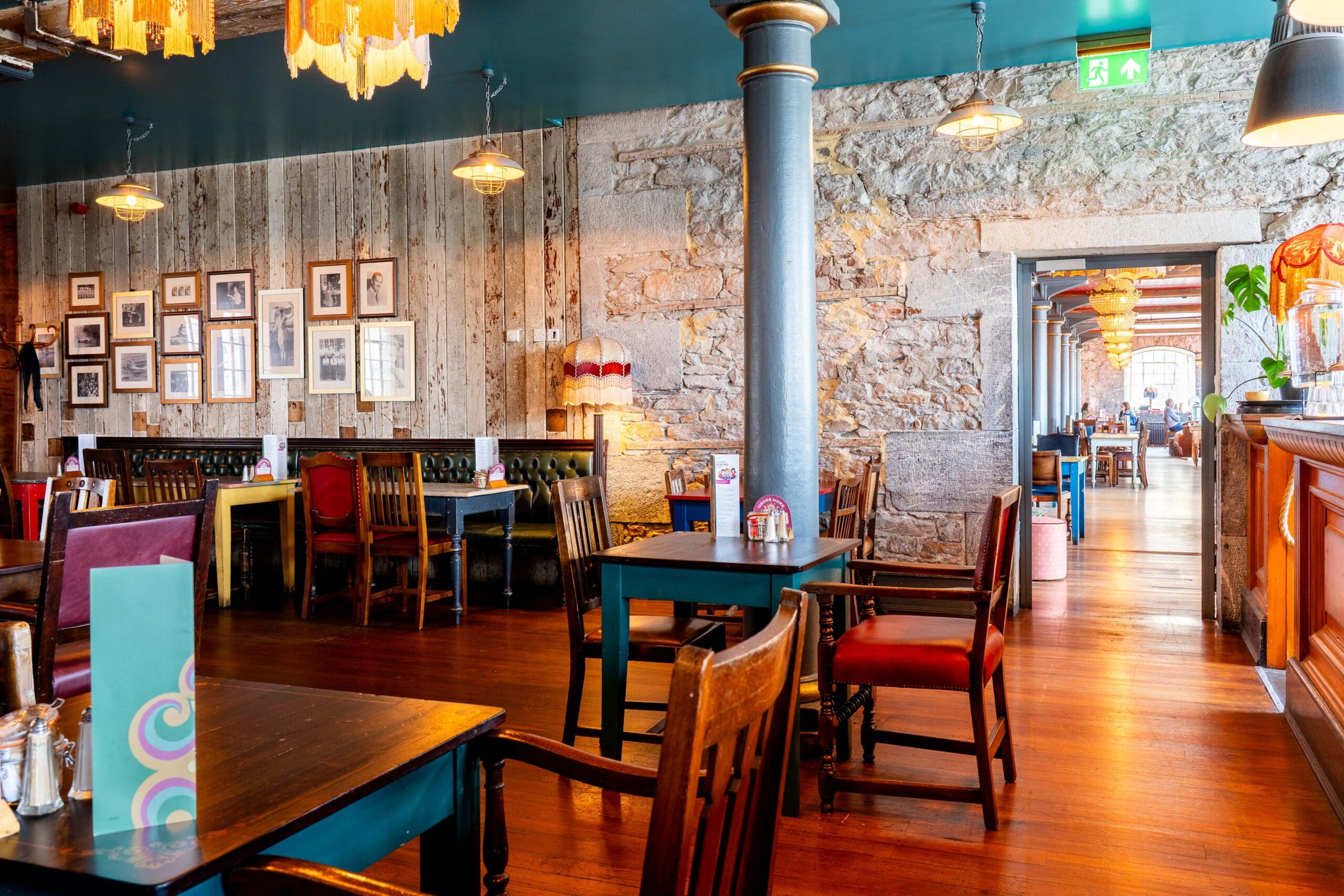 A cozy restaurant interior with wooden floors, rustic stone walls, eclectic chairs, framed photos on the wall, and warm lighting. A hallway leads to more seating in the background.