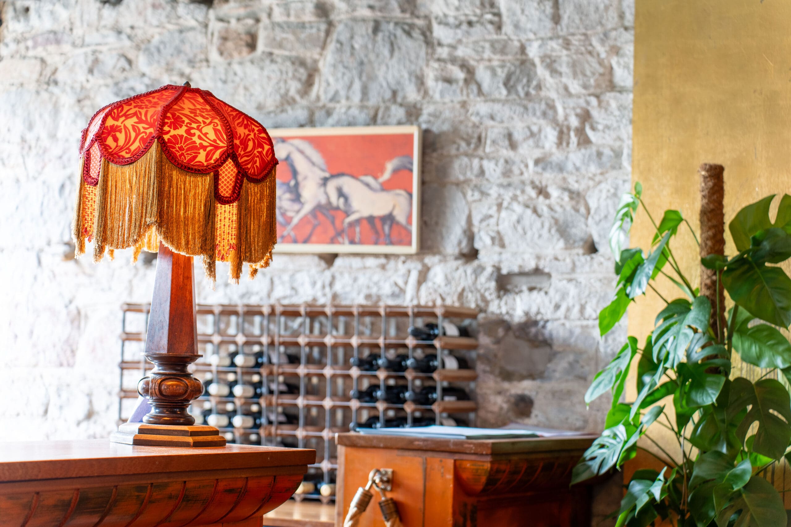 A vintage red and gold fringed lamp sits on a wooden counter in front of a stone wall, with a plant, wine rack, and framed horse artwork in the background.
